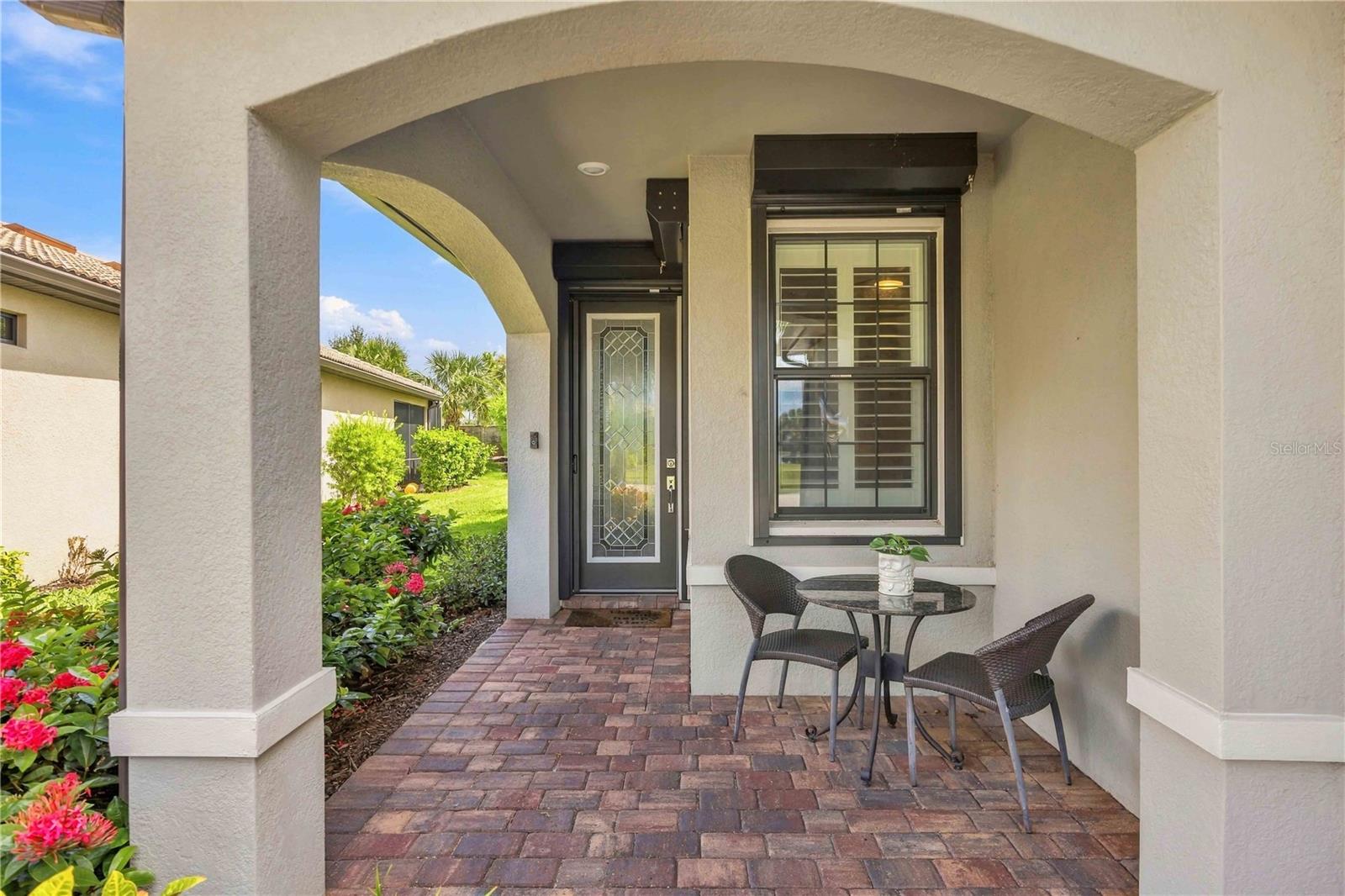 Covered front porch with a stucco arch, brick pavers, glass-paneled door, and small seating area.