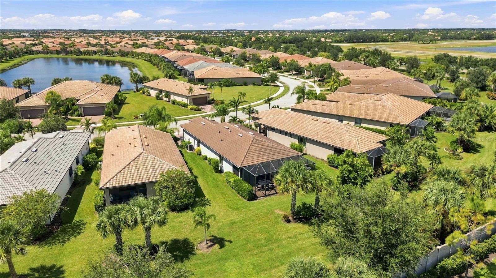 Aerial view of homes with tile roofs and palm-lined lawns beside a neighborhood pond.