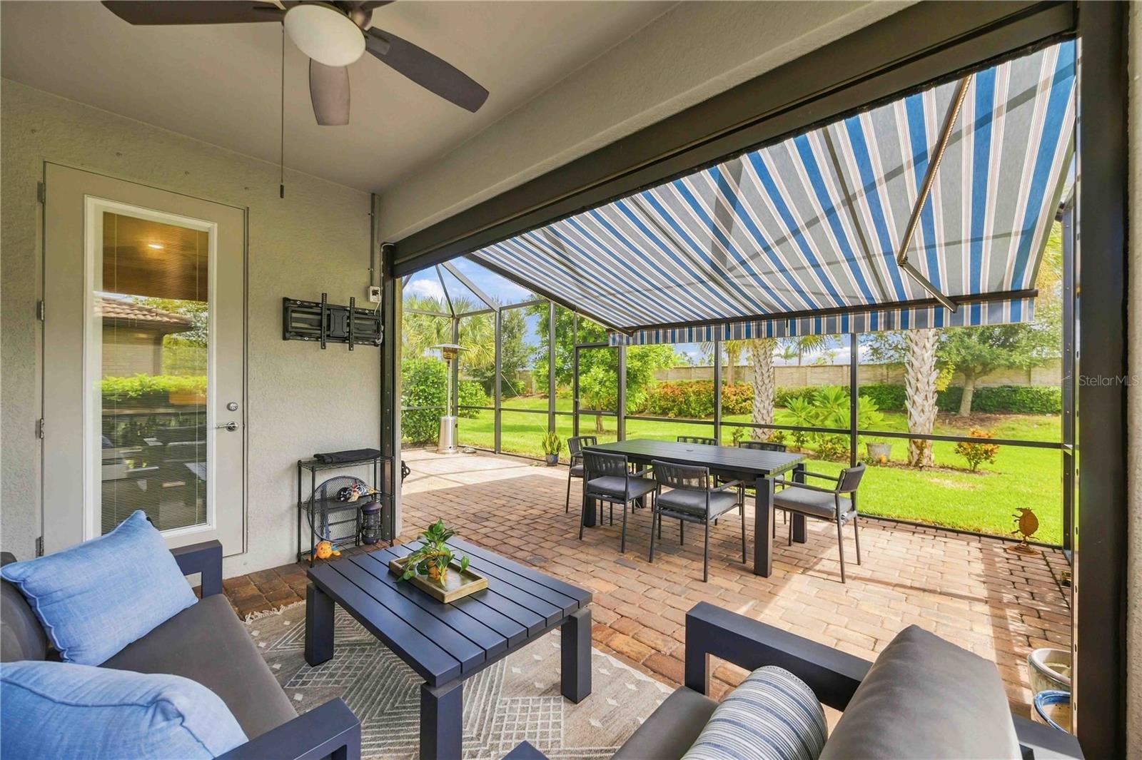 Screened patio with brick pavers, outdoor seating, and a striped retractable awning overlooking a palm-lined yard.