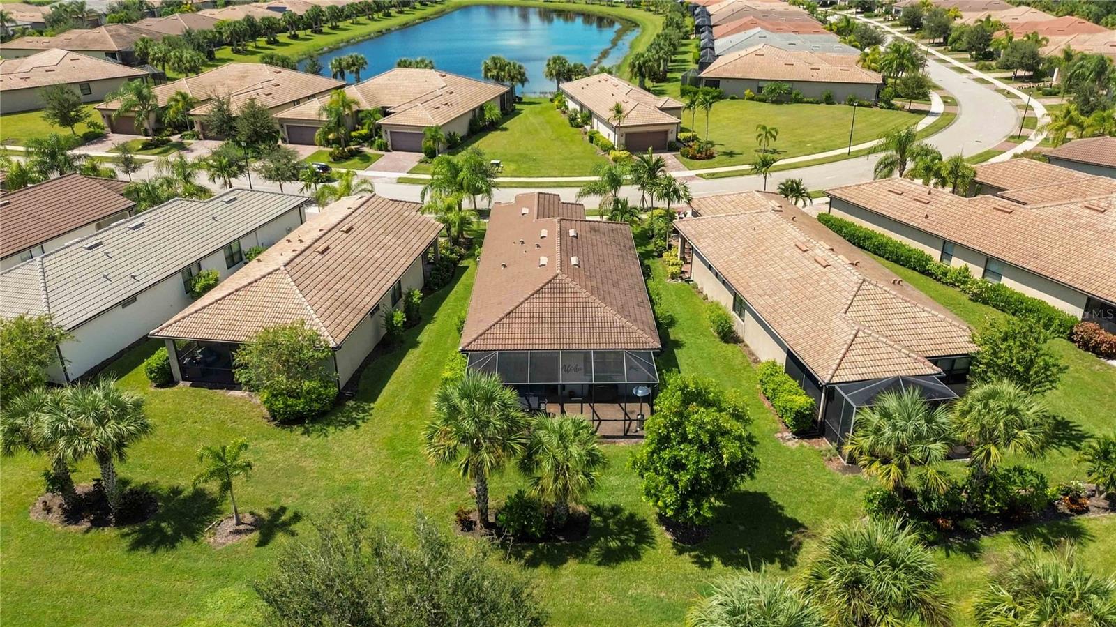 Aerial view of a tile-roof home with a screened lanai, palms, and a lake nearby.