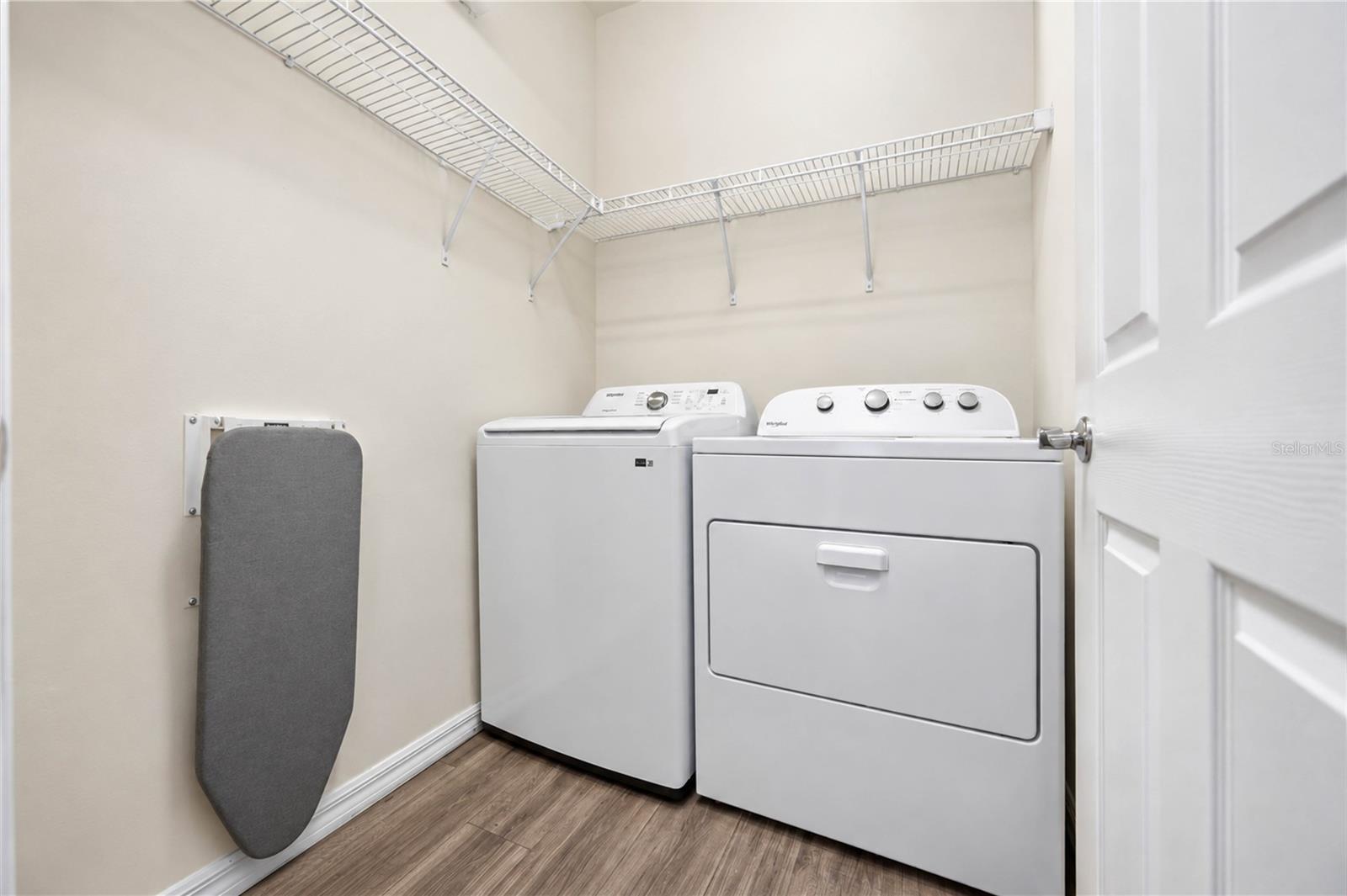 Laundry room with a washer and dryer, wraparound wire shelving, and a wall-mounted ironing board.