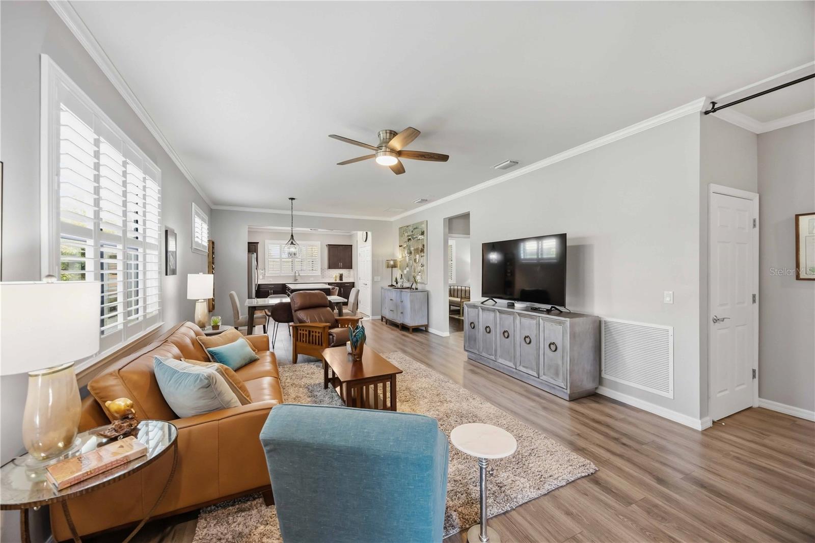Open-plan living room with tan leather sofa, plantation shutters, and dining area leading to the kitchen.