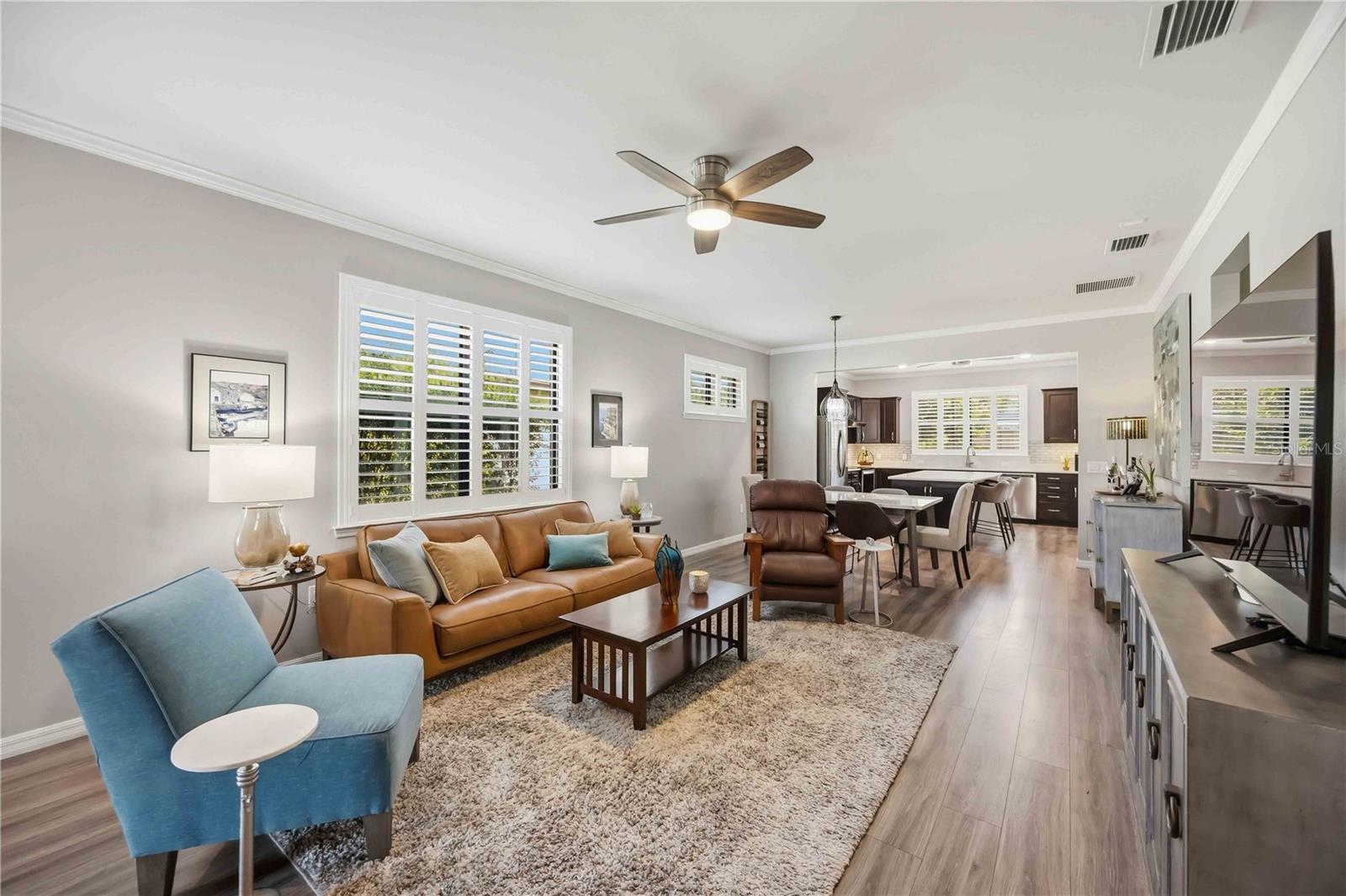 Open-plan living room and kitchen with plantation shutters, a tan sofa, and wood-look floors.