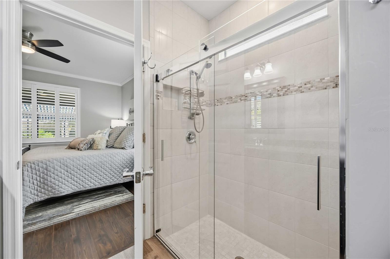 Tiled walk-in shower with glass doors, viewed beside a bedroom with plantation shutters.