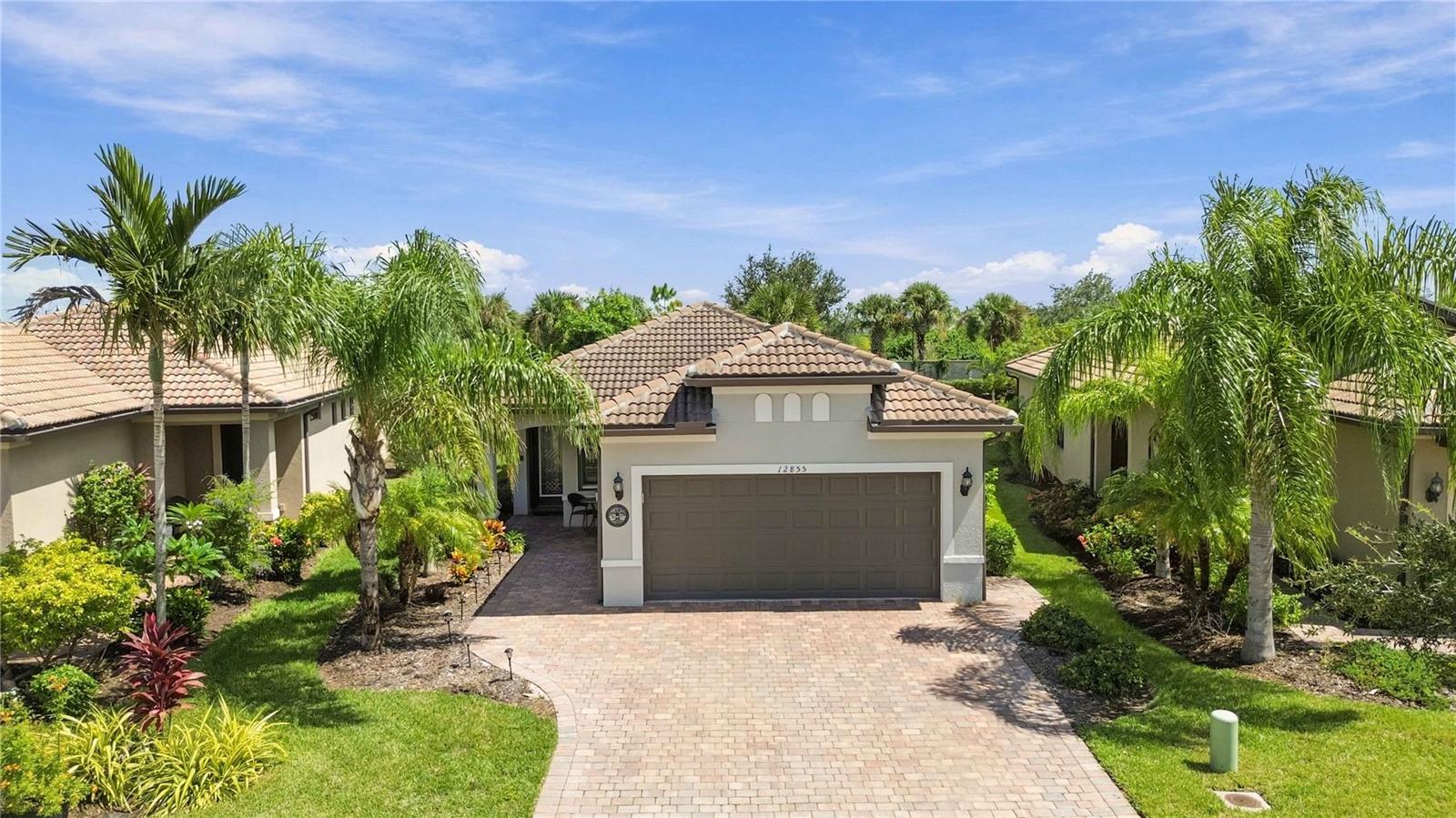 Single-story home with a tiled roof, two-car garage, paver driveway, and tropical landscaping.