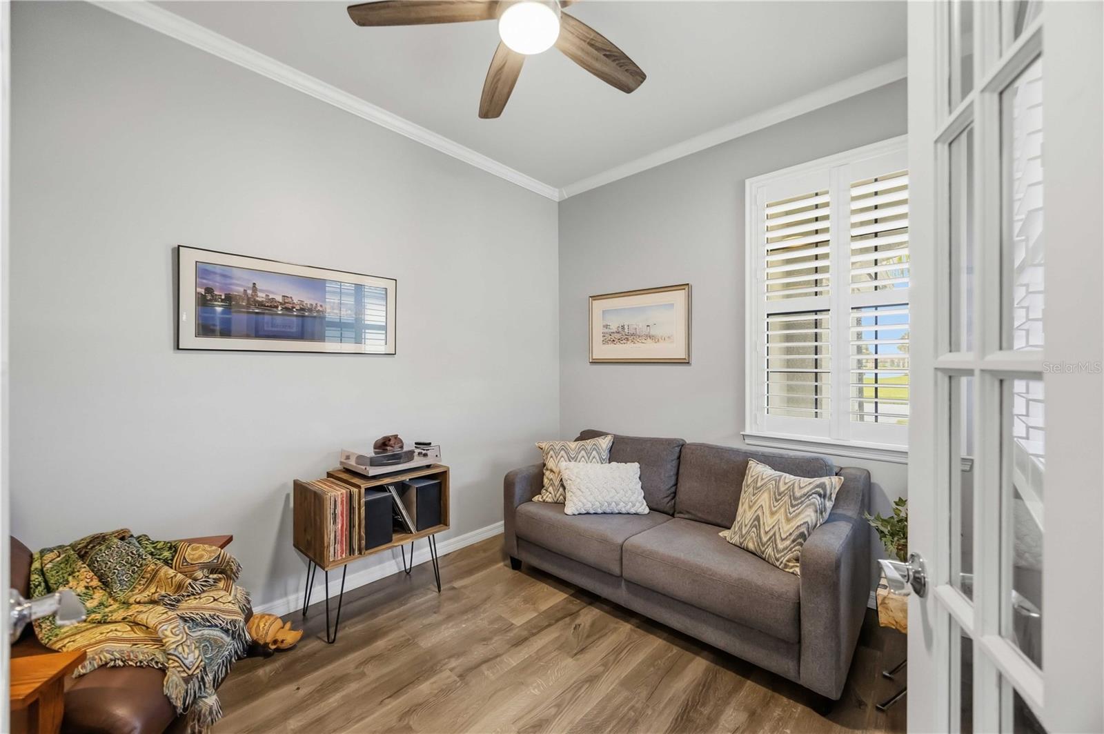 Living room with a gray sofa, shuttered window, wood-look floors, and ceiling fan.