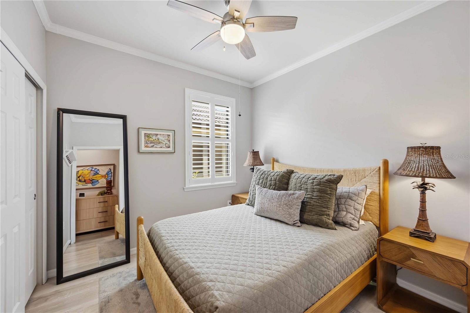 Furnished bedroom with a light wood bed, gray quilted bedding, plantation shutters, and a large leaning mirror.