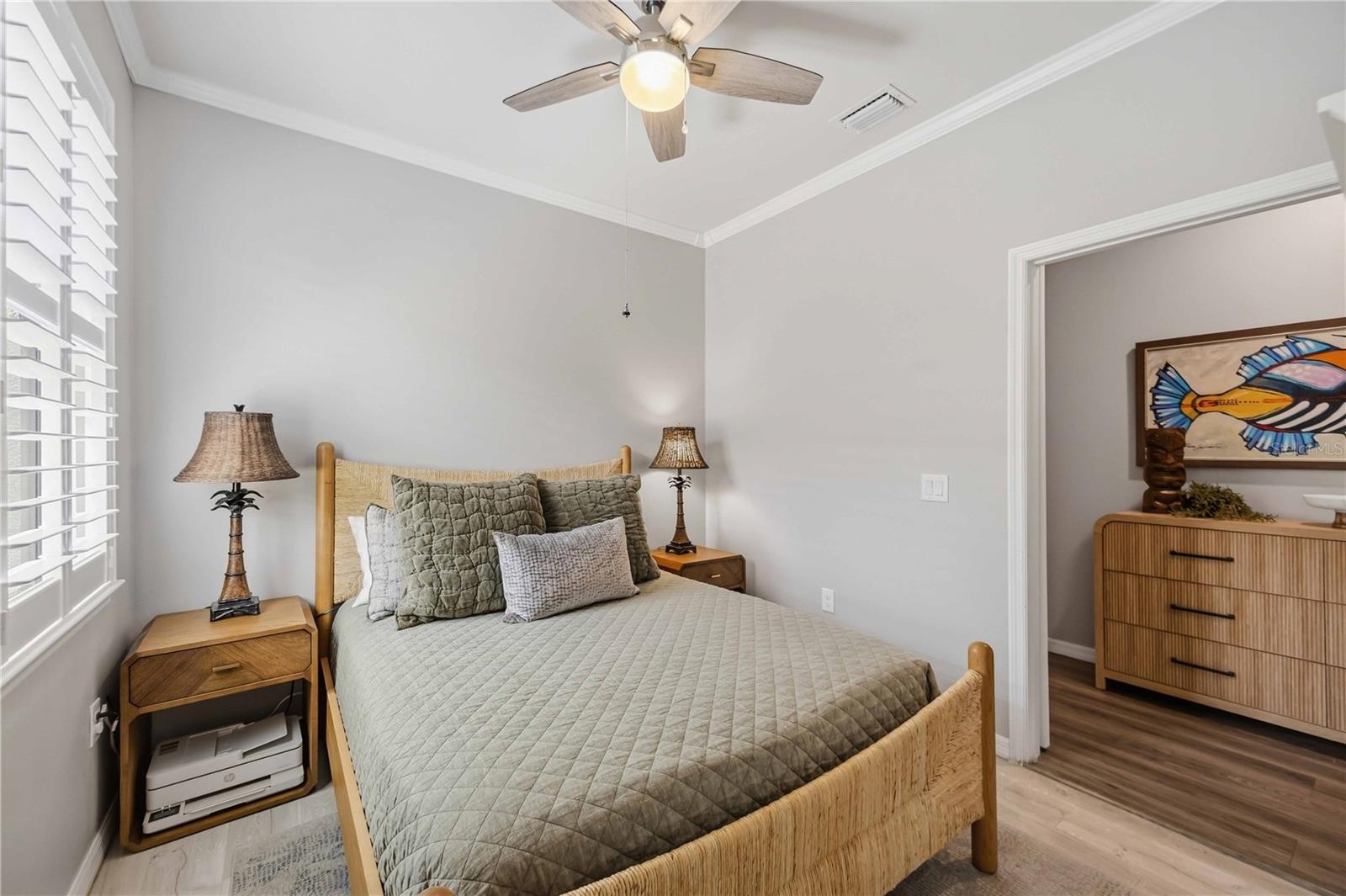 Bedroom with a rattan bed, gray quilt, plantation shutters, and colorful fish artwork visible through the doorway.