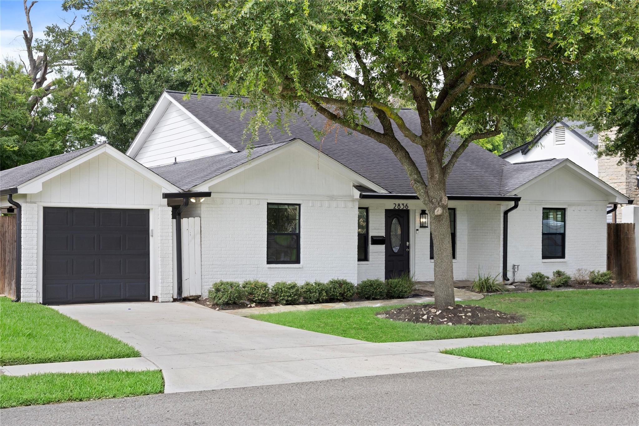 White-brick ranch-style home with a dark garage door beneath a large shade tree.