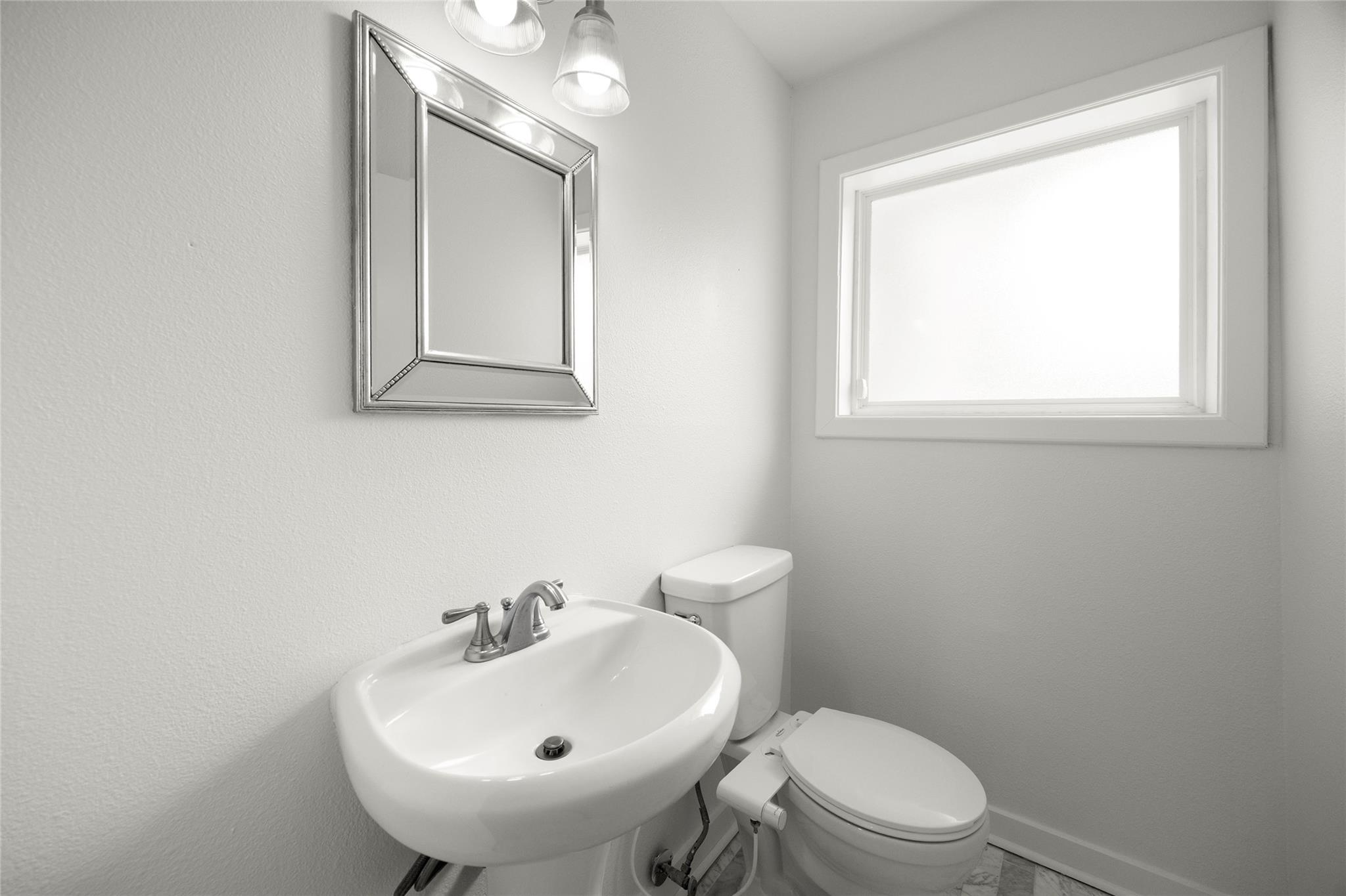 Bathroom with a white pedestal sink, toilet, framed mirror, and high window.
