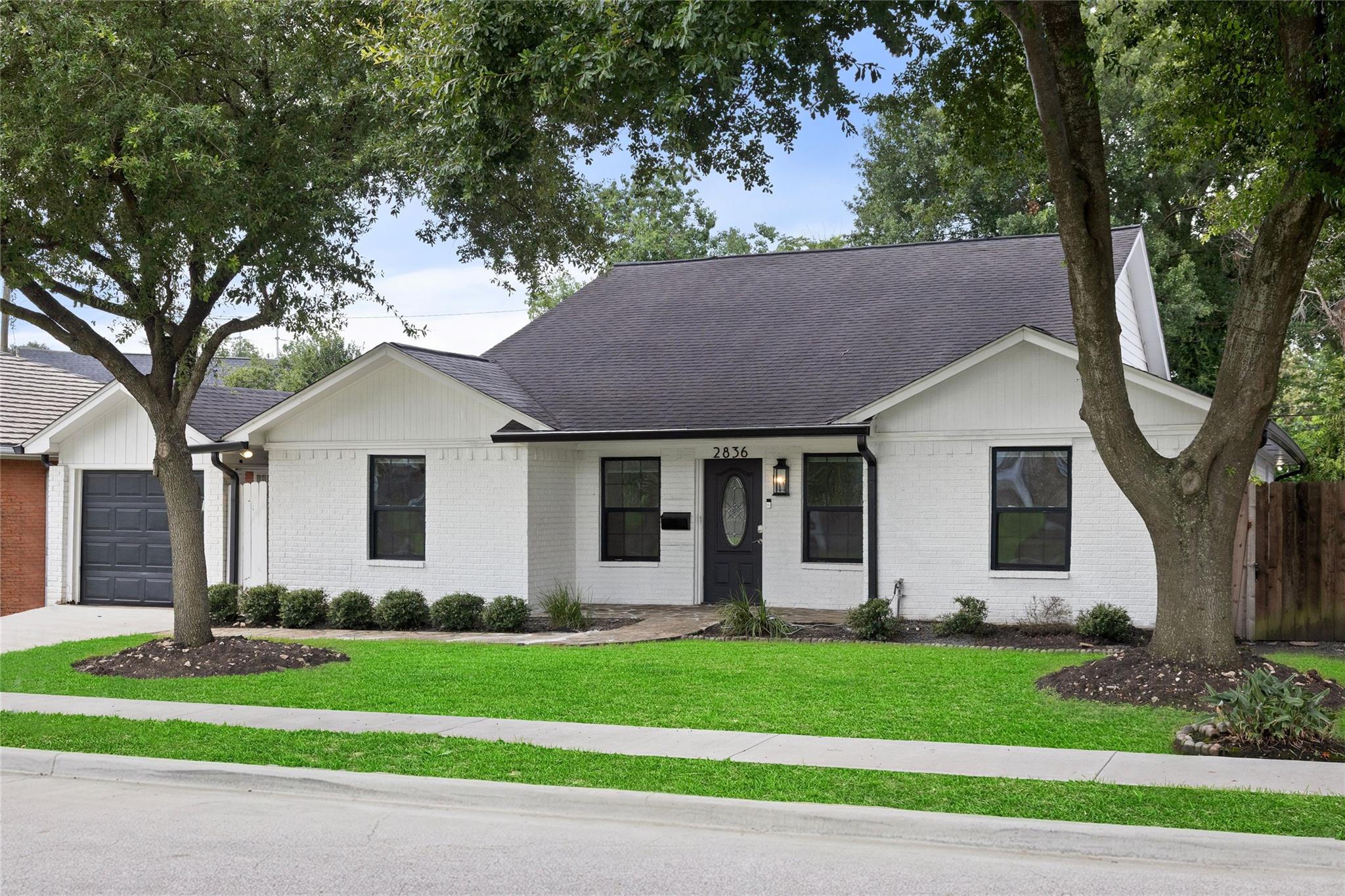White brick house with dark trim and attached garage, framed by mature trees and a green lawn.