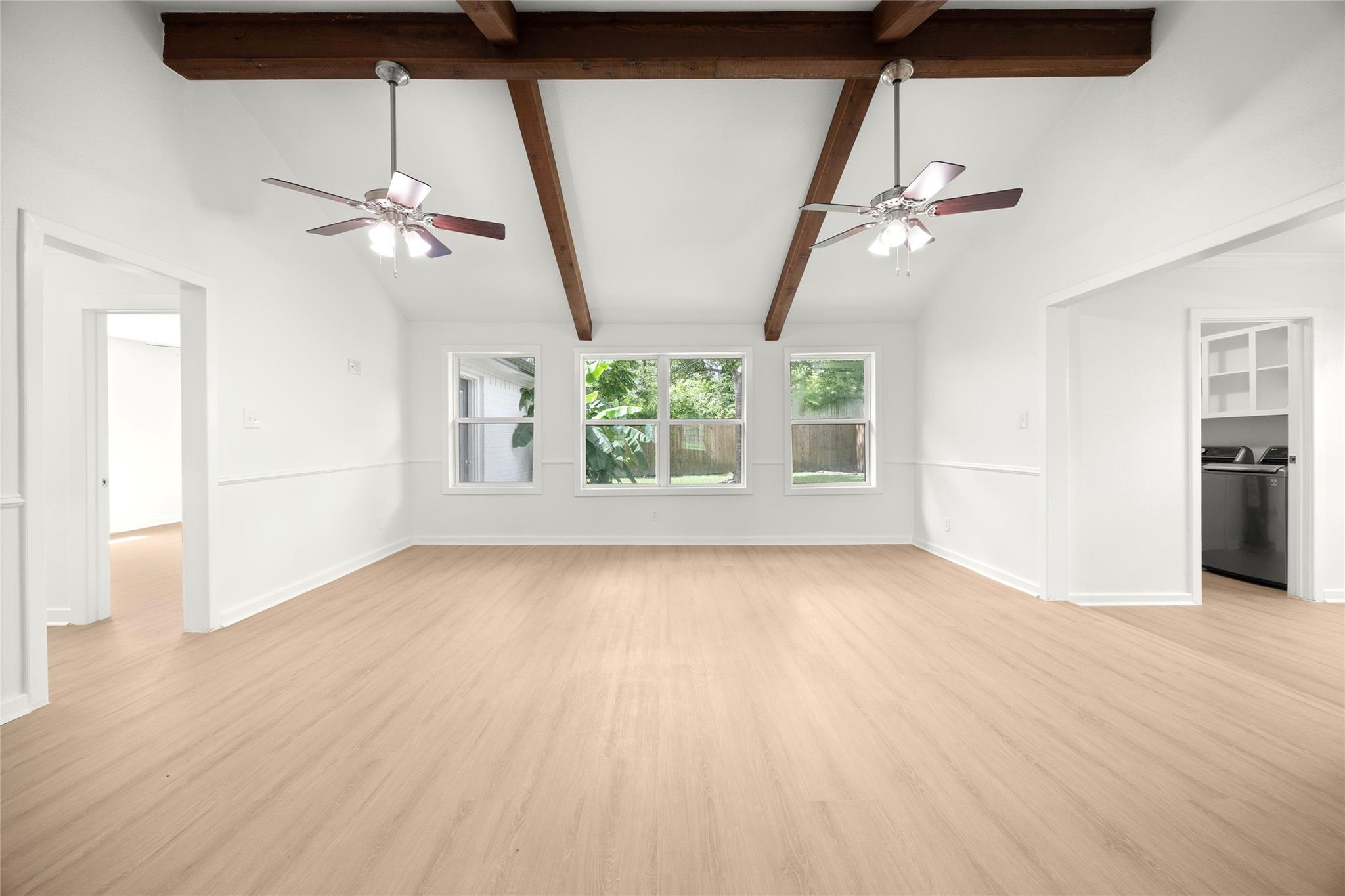 Empty living room with exposed wood ceiling beams, two ceiling fans, and windows overlooking greenery.