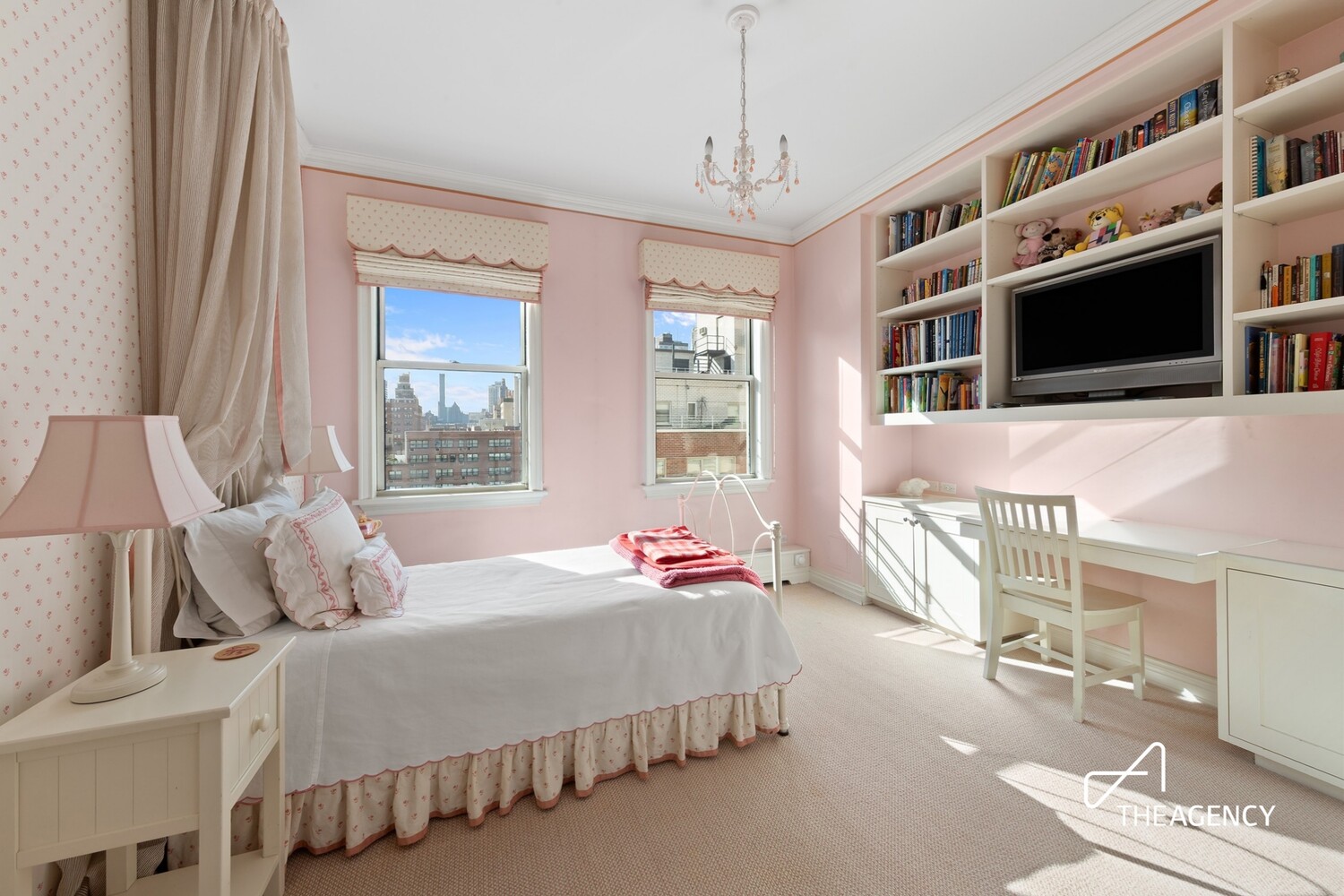 Pink bedroom with white bed, built-in bookcases and desk, and city views through two windows.