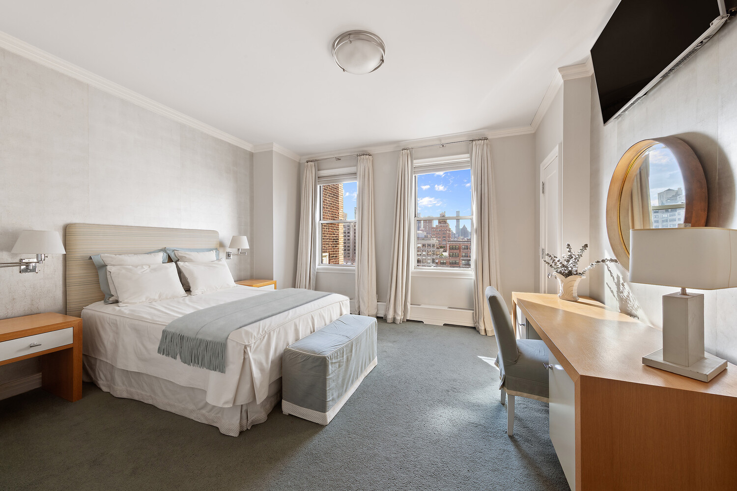Furnished bedroom with a large bed, wood desk, and city views through two windows.