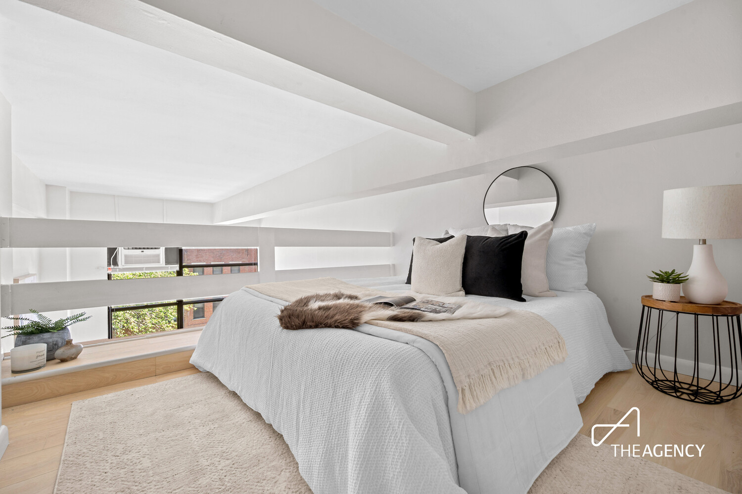 Staged bedroom with a white bed, neutral pillows, a round mirror, and a low window with horizontal shades.