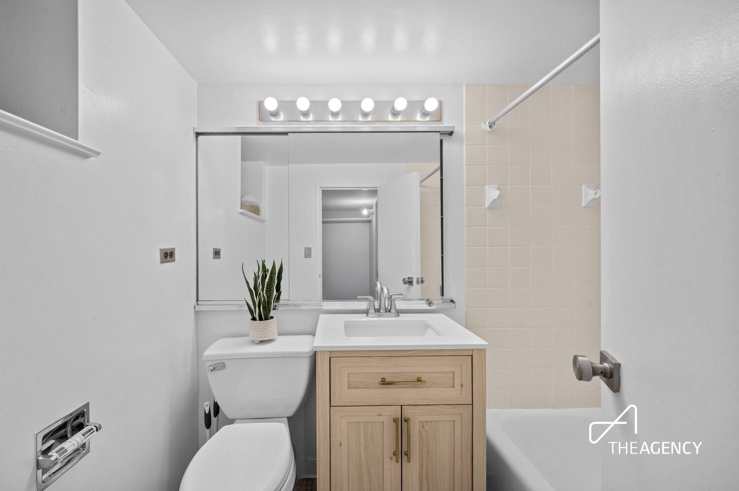 Bathroom with a light wood vanity, mirrored cabinet, and white tub with tiled surround.