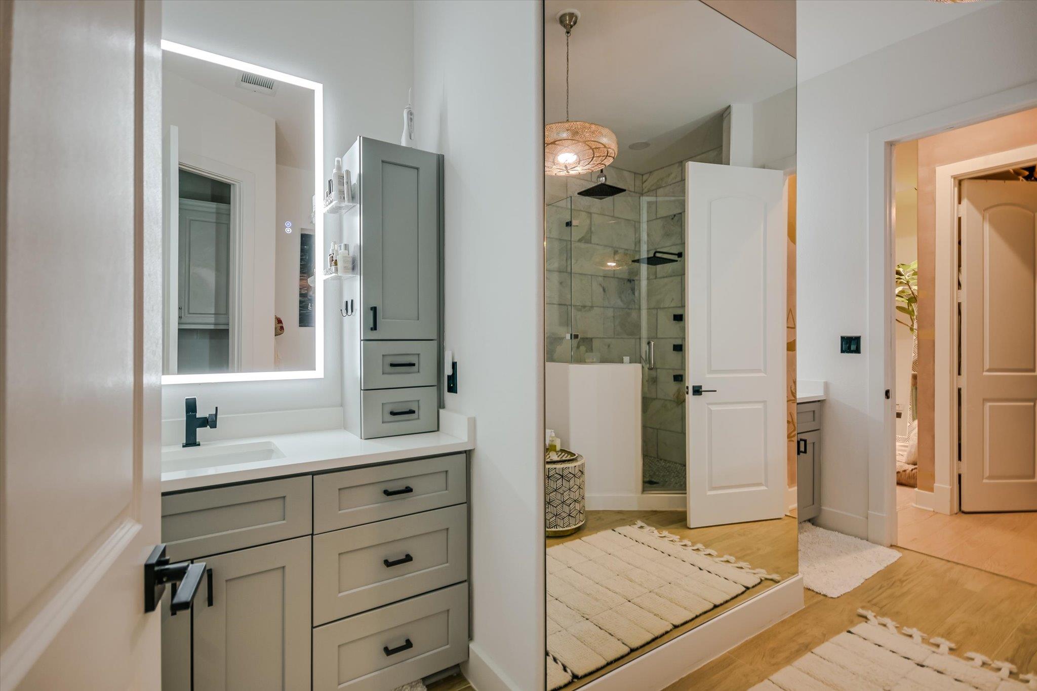 Modern bathroom with gray vanity, illuminated mirror, and glass-enclosed marble-look shower.