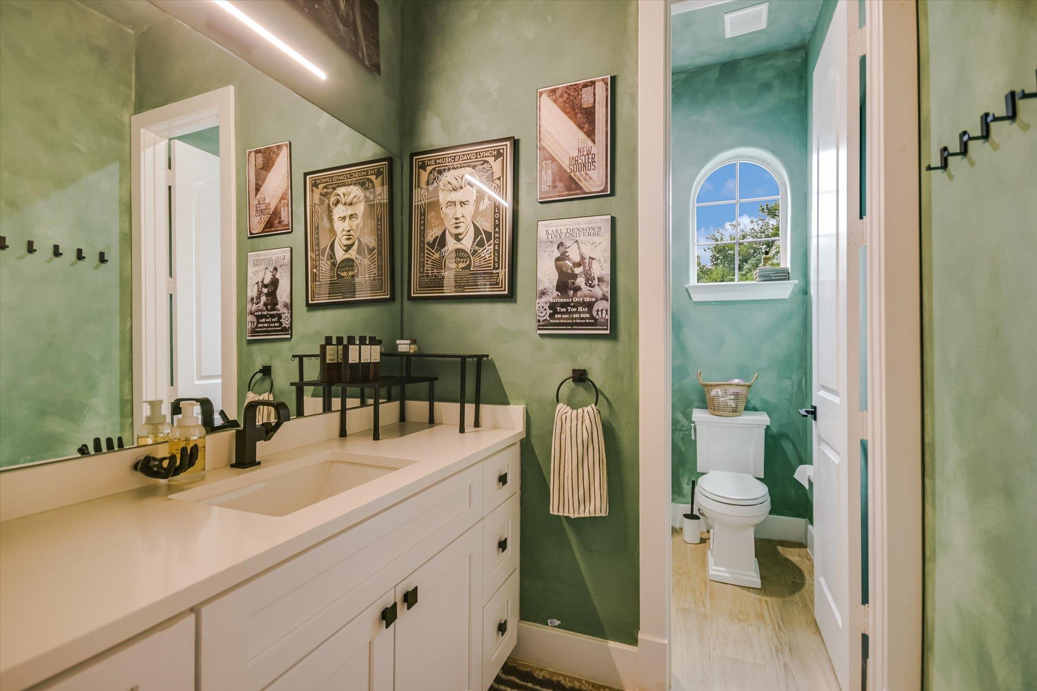 Bathroom with a long white vanity and green walls; an arched window sits above the toilet in the adjoining room.