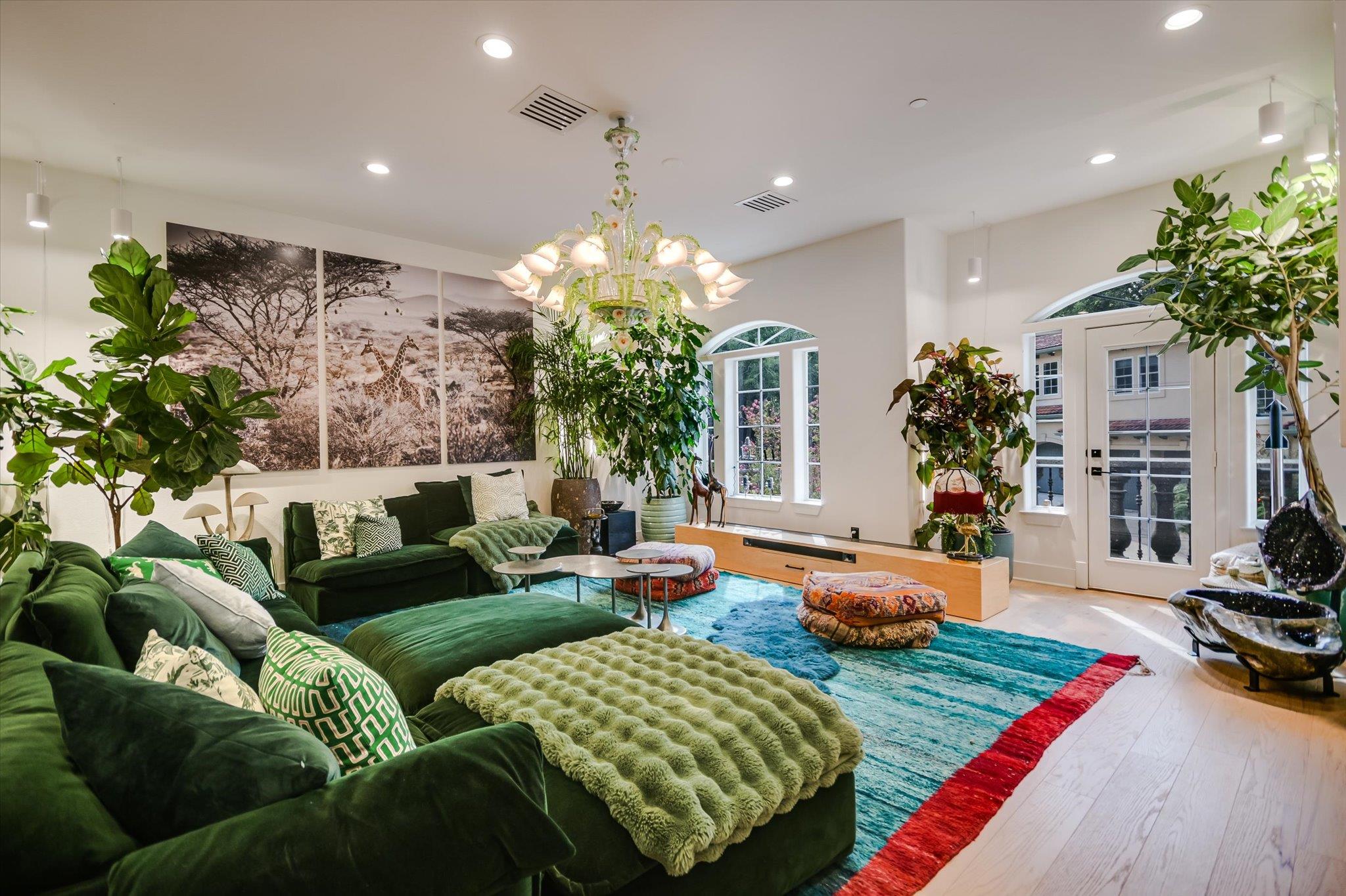Living room with green sectional sofas, a turquoise rug, large windows, plants, and a floral chandelier.