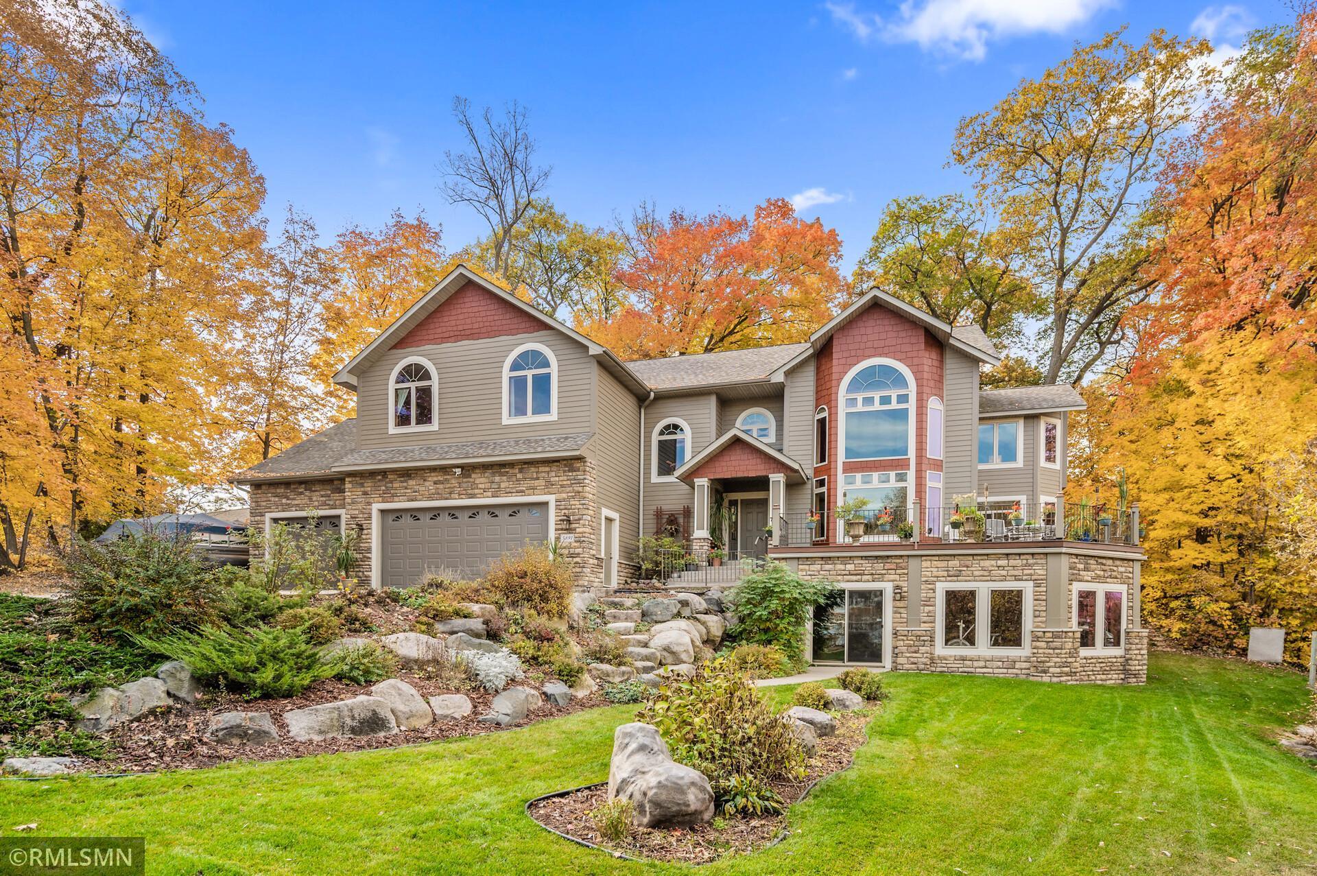 Two-story taupe home with stonework, raised patio, landscaped rock garden, and colorful autumn trees.