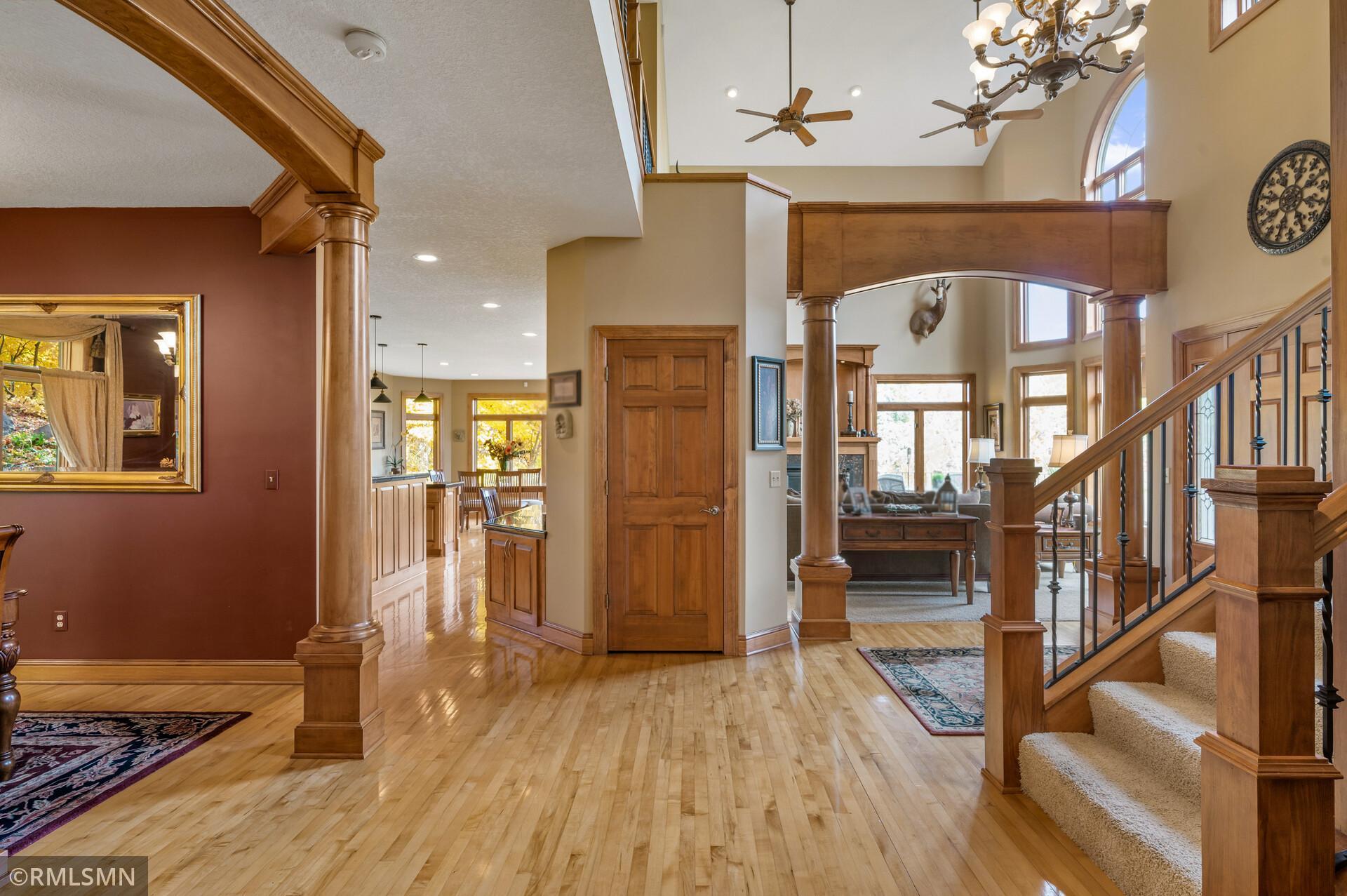 Foyer with glossy hardwood floors, wood columns, and a carpeted staircase beside a tall arched window.