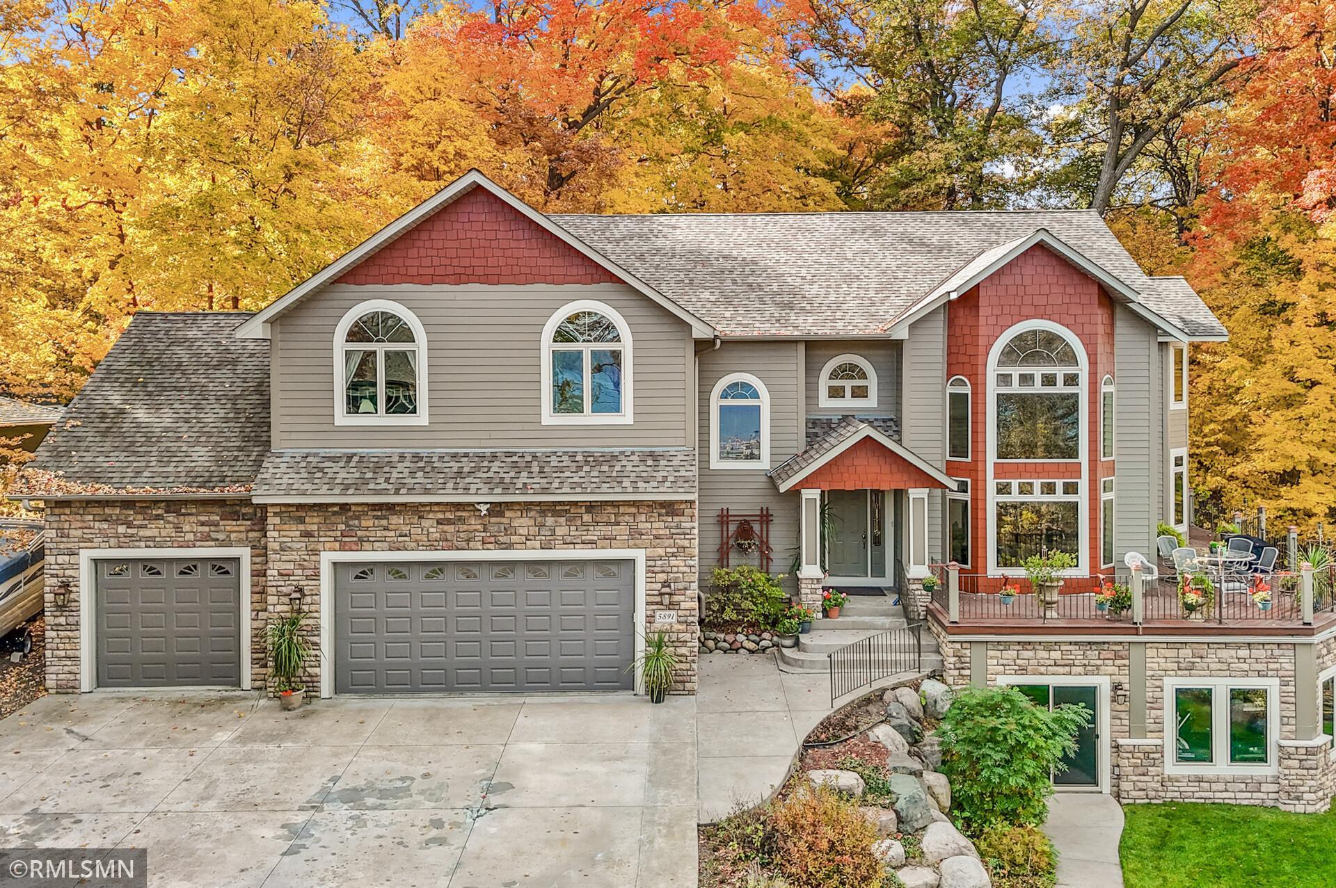 House exterior with gray siding, red accents, stonework, and a raised patio amid autumn trees.