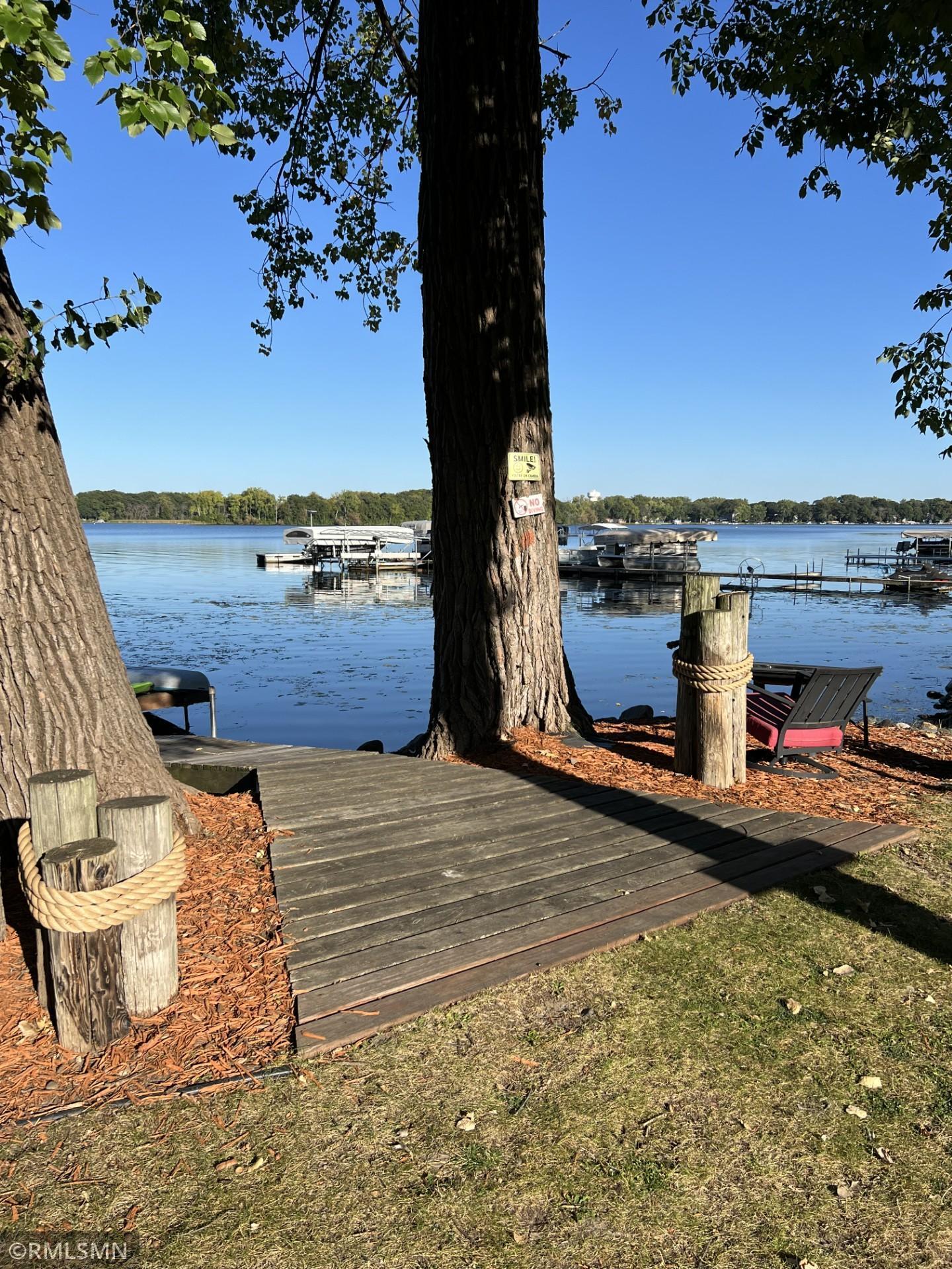 Wooden lakeside dock beneath mature trees, with rope-wrapped posts and boats across the water.