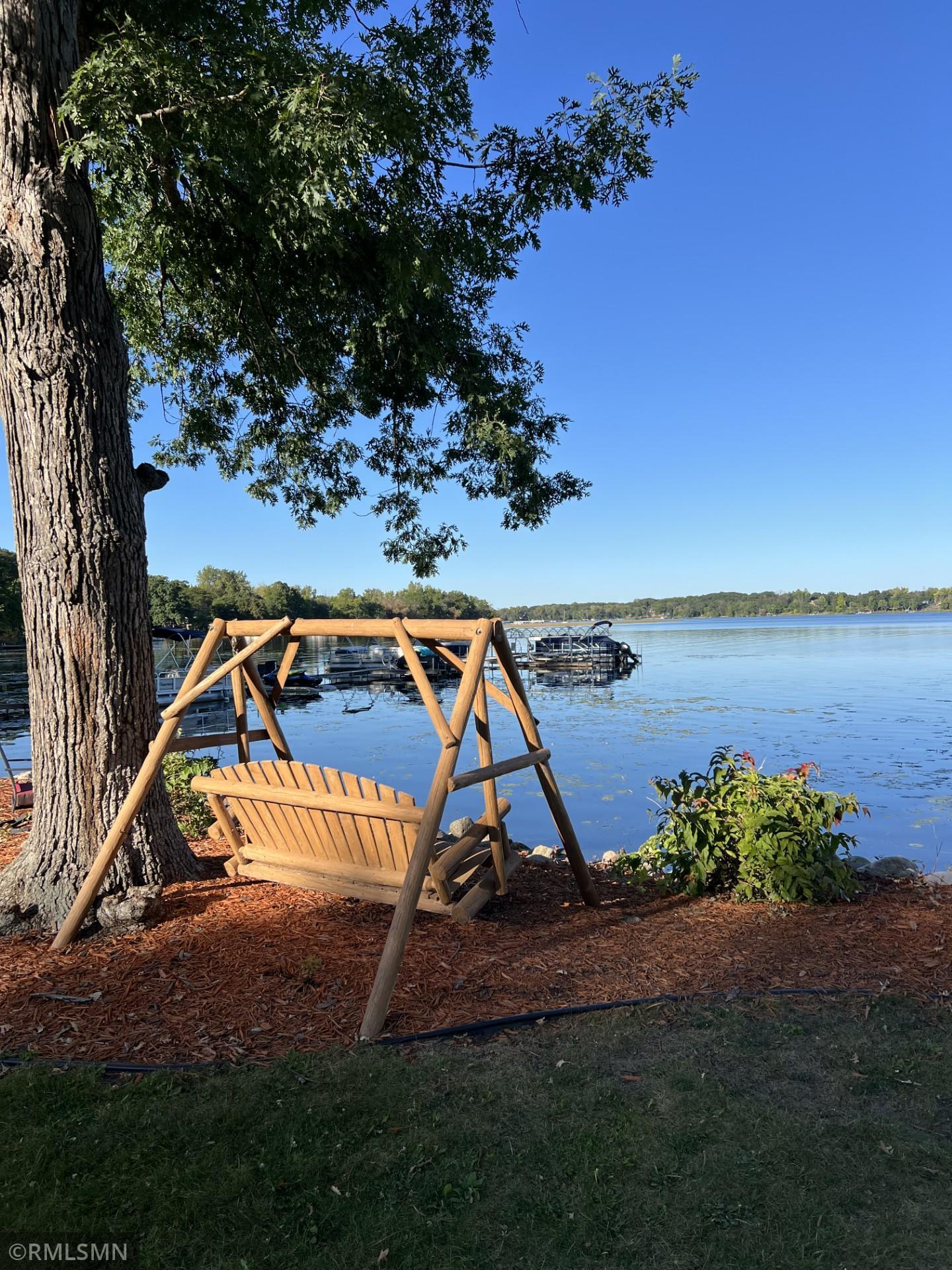 Wooden porch swing beneath a large tree overlooking a lake, boats, and docks.