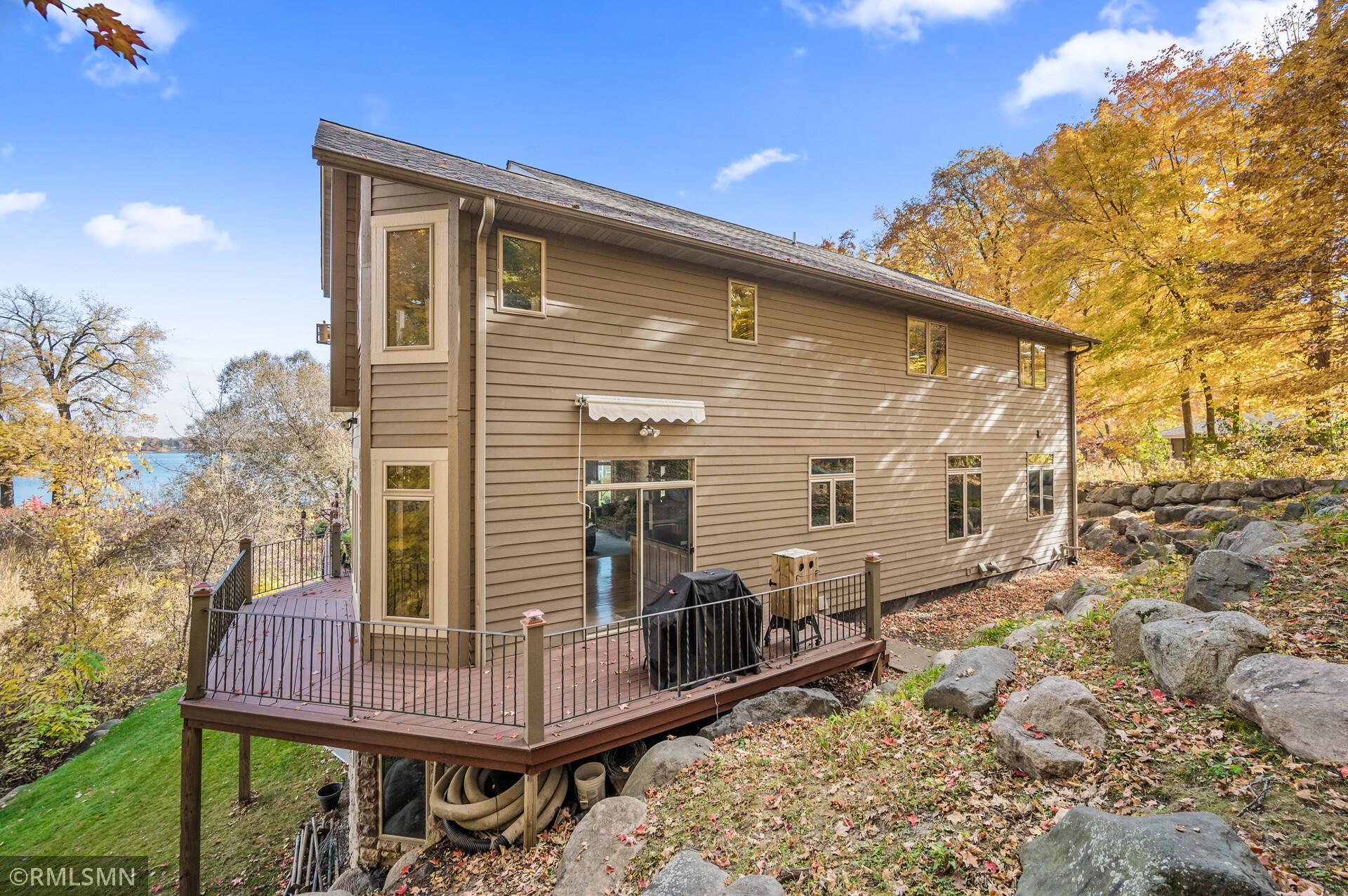 Tan two-story house with a raised deck among autumn trees and rocks, with lake water visible through the trees.