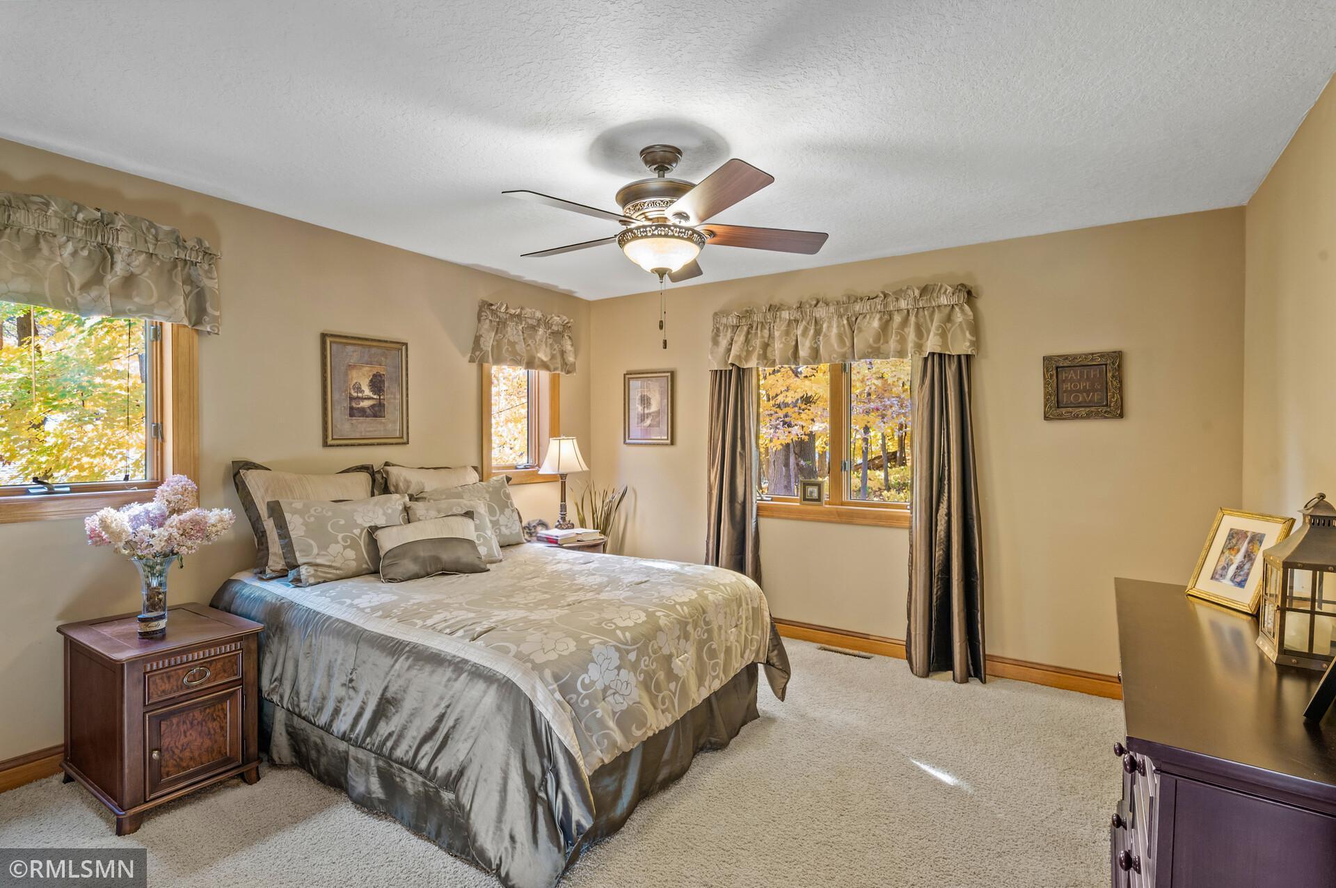 Furnished bedroom with a large patterned bed, ceiling fan, and windows overlooking autumn foliage.