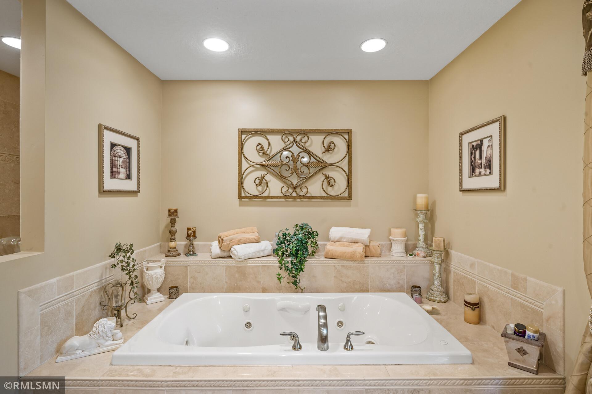 Bathroom soaking tub with beige tile surround, folded towels, plants, and ornate metal wall art.