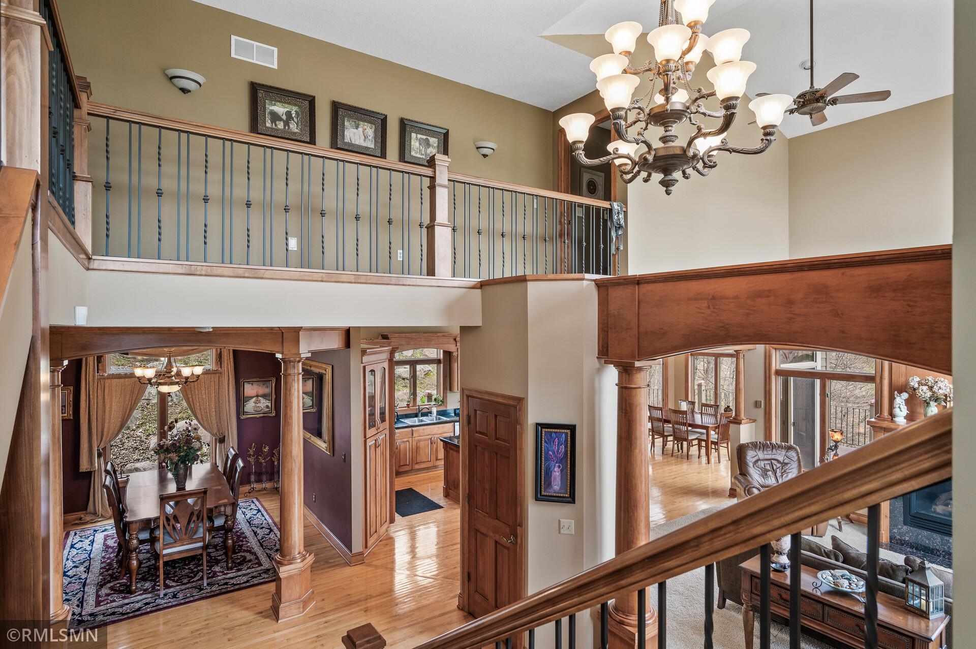 Two-story interior with a wood-railed balcony, ornate chandelier, and views into dining and living areas.