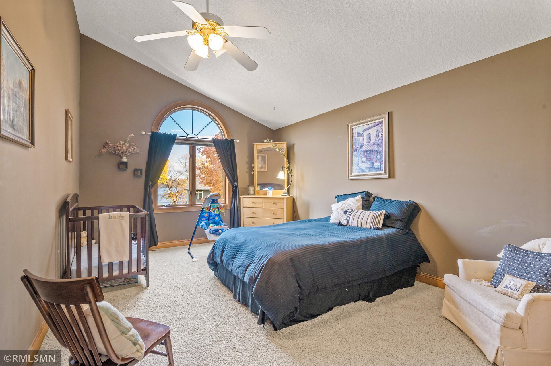 Bedroom with vaulted ceiling, arched window overlooking water, large bed, and wooden crib.