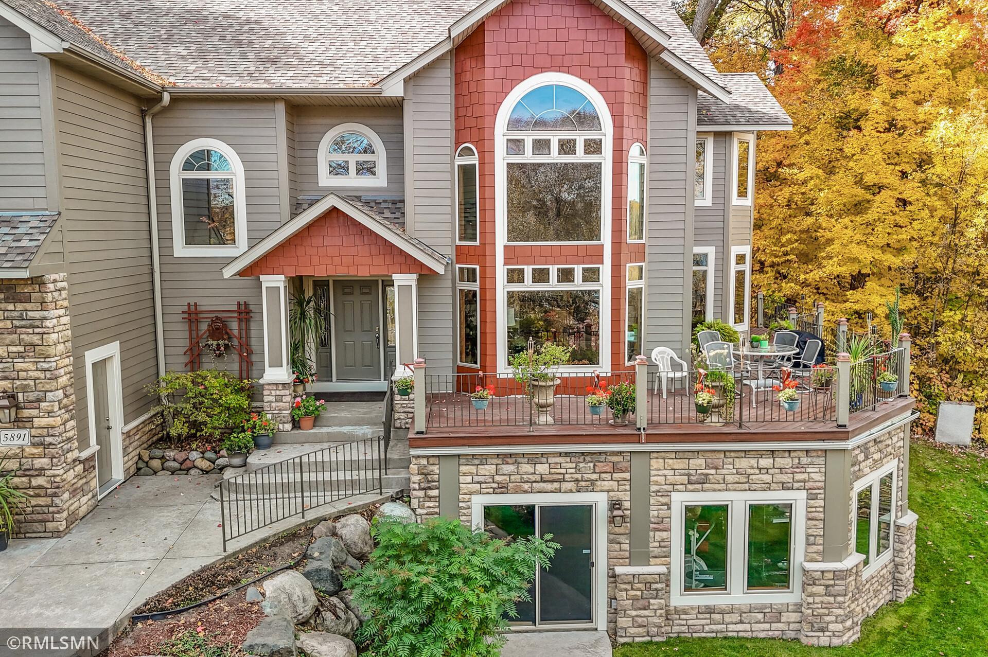 Home exterior with red shingle accents, tall arched windows, and a furnished deck amid autumn trees.