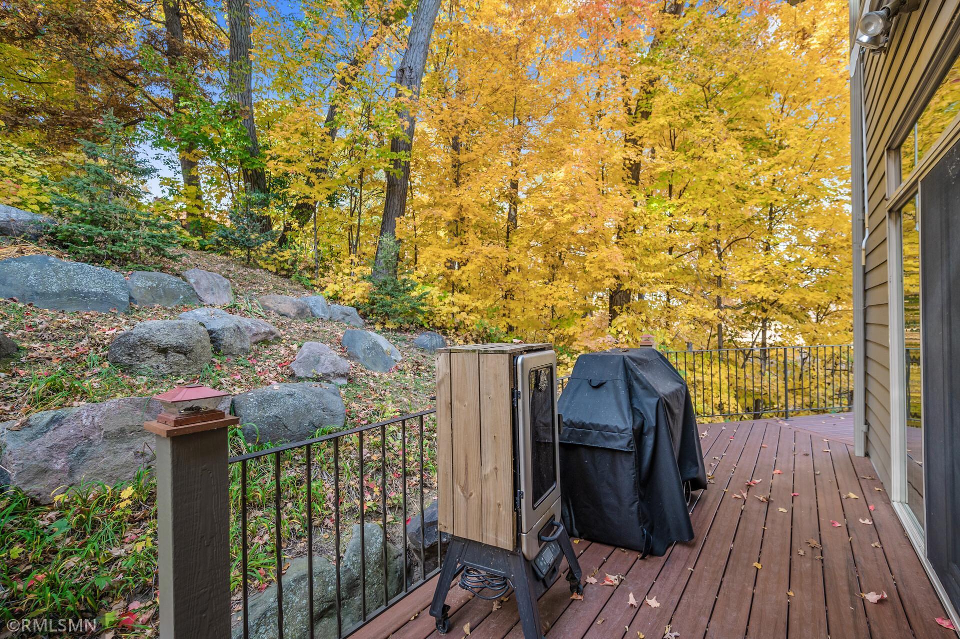 Wooden deck with a covered grill overlooking a rocky, tree-filled slope in autumn.
