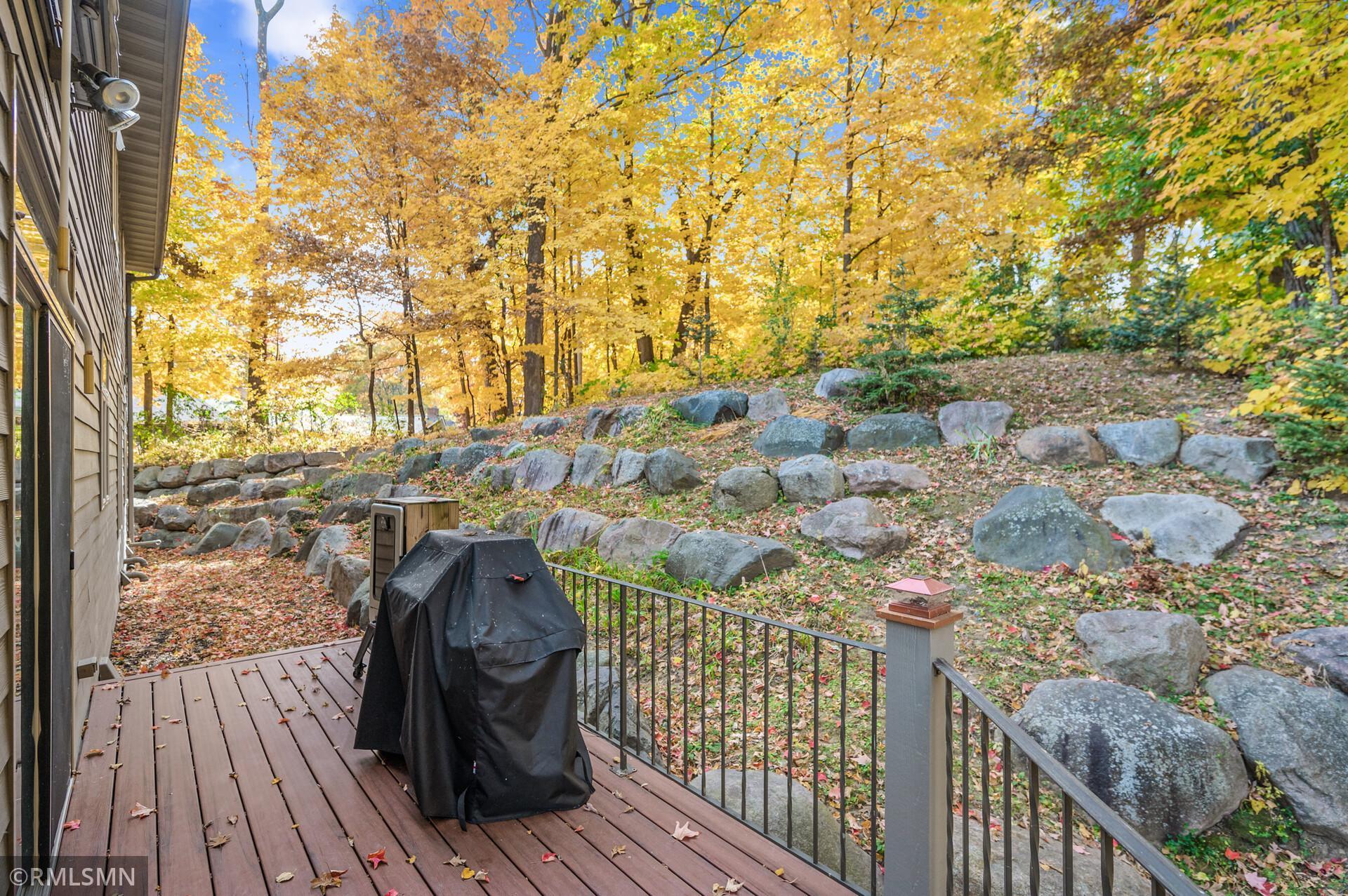 Wooden deck overlooking a sloped, rock-lined backyard with golden autumn trees and a covered grill.