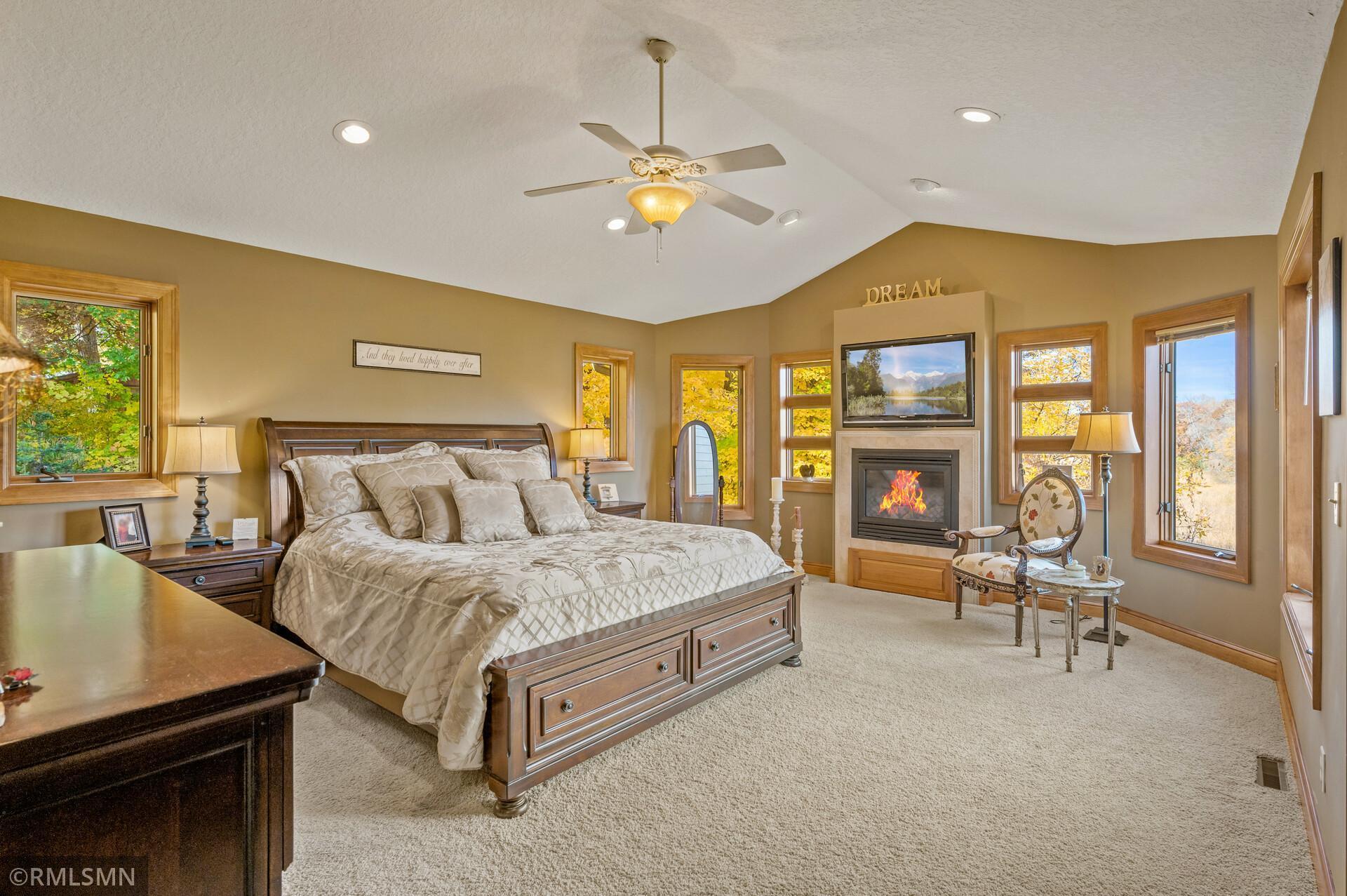 Furnished bedroom with a wooden bed, fireplace, and windows overlooking autumn foliage.