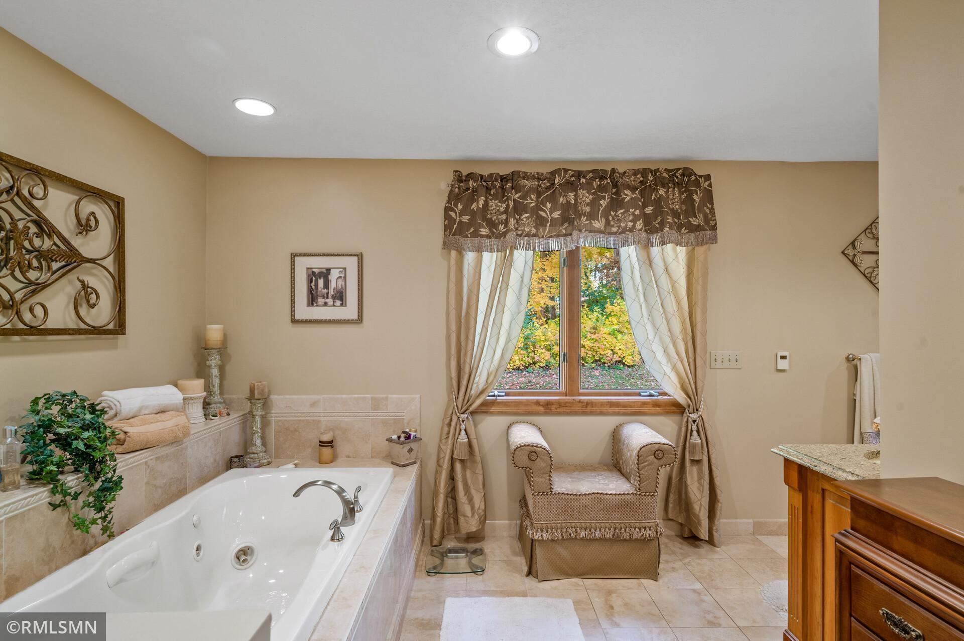Bathroom with a corner soaking tub and cushioned armchair beside a curtained window.