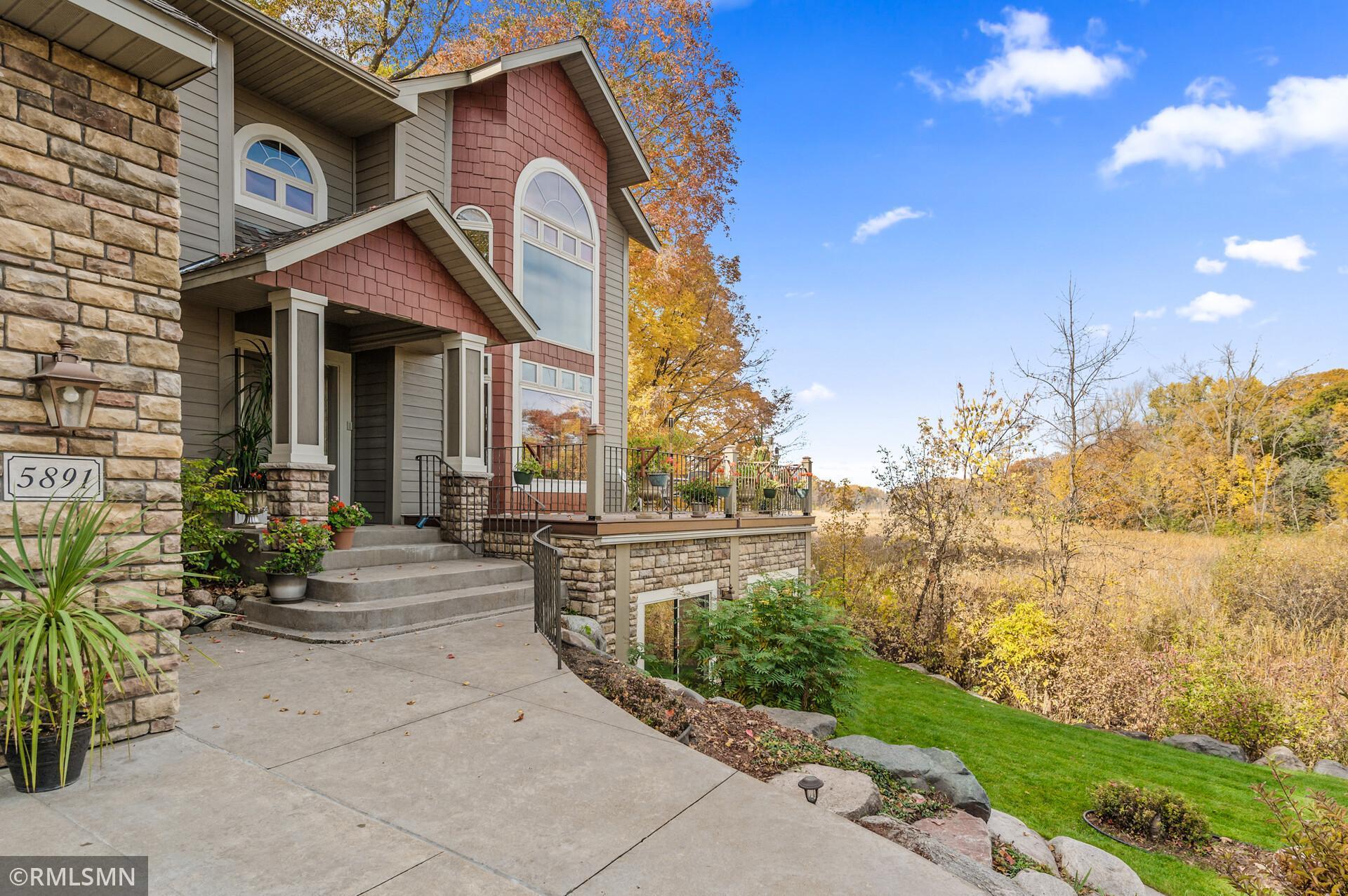 Home with gray siding, red accents, stonework, and a raised porch beside autumn trees.