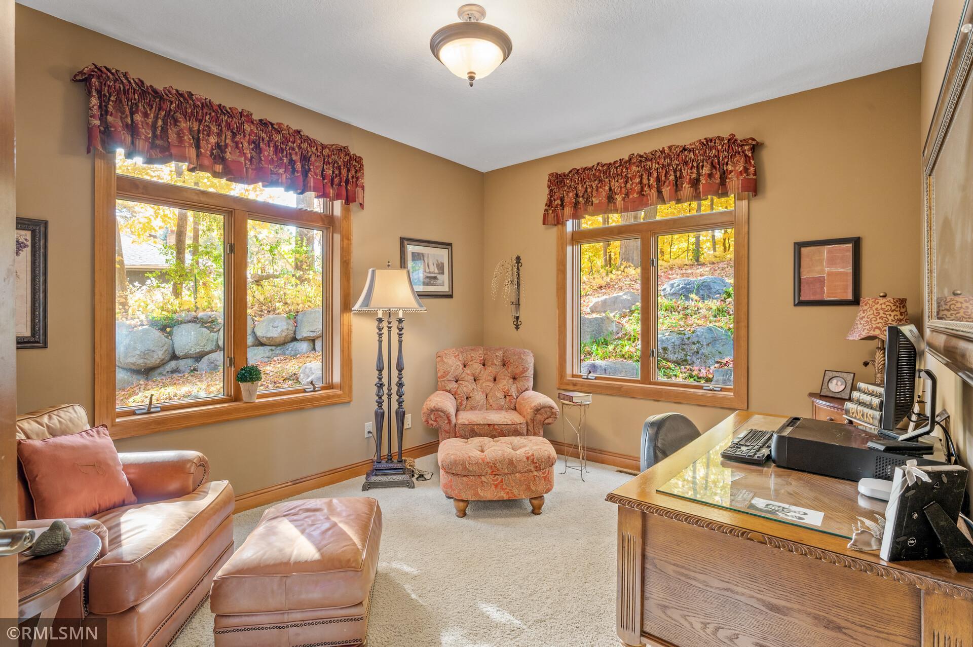 Furnished home office with a carved wood desk, leather seating, and windows overlooking autumn foliage.