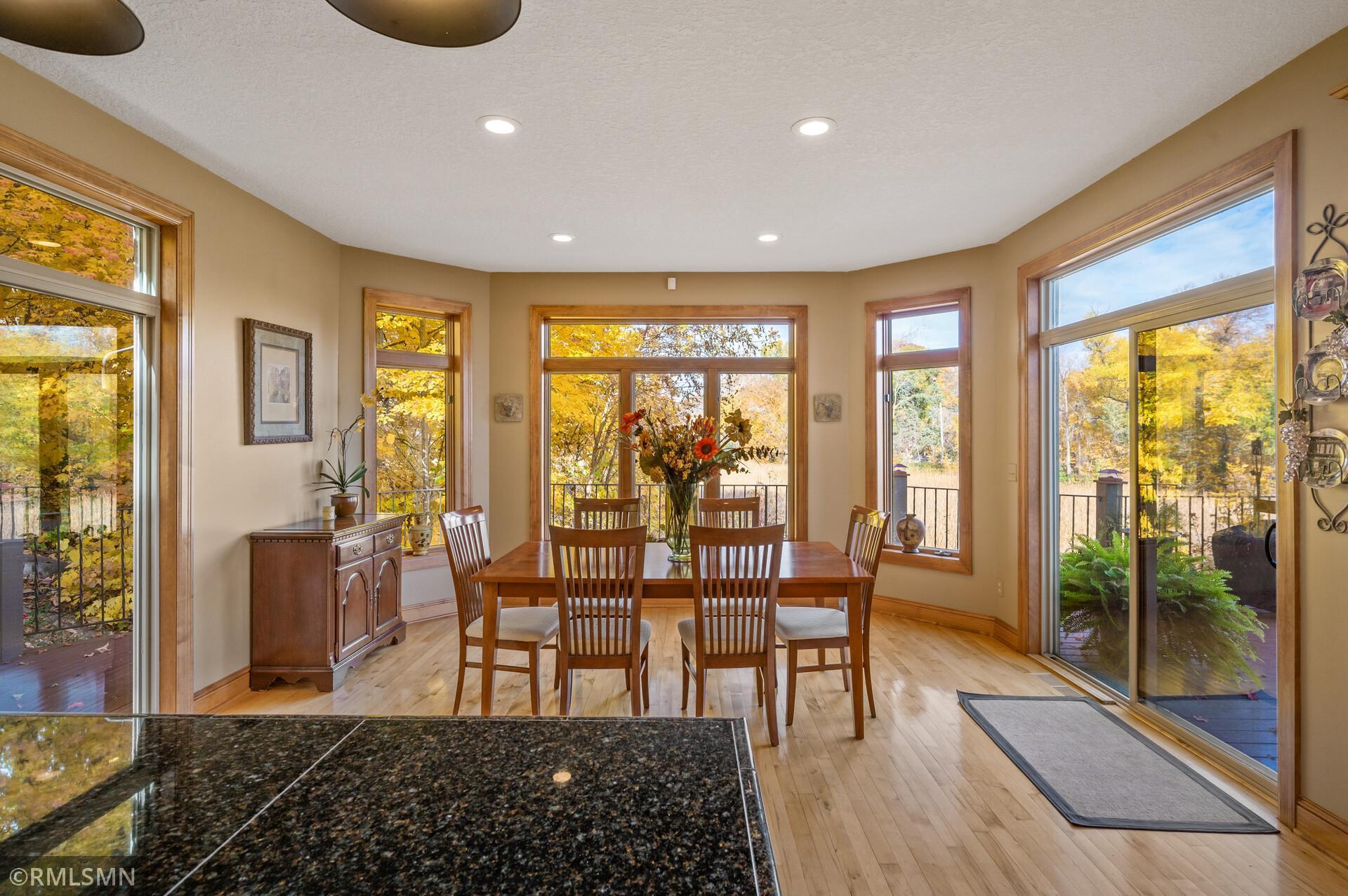 Dining room with a wooden table and chairs, surrounded by large windows overlooking autumn foliage.
