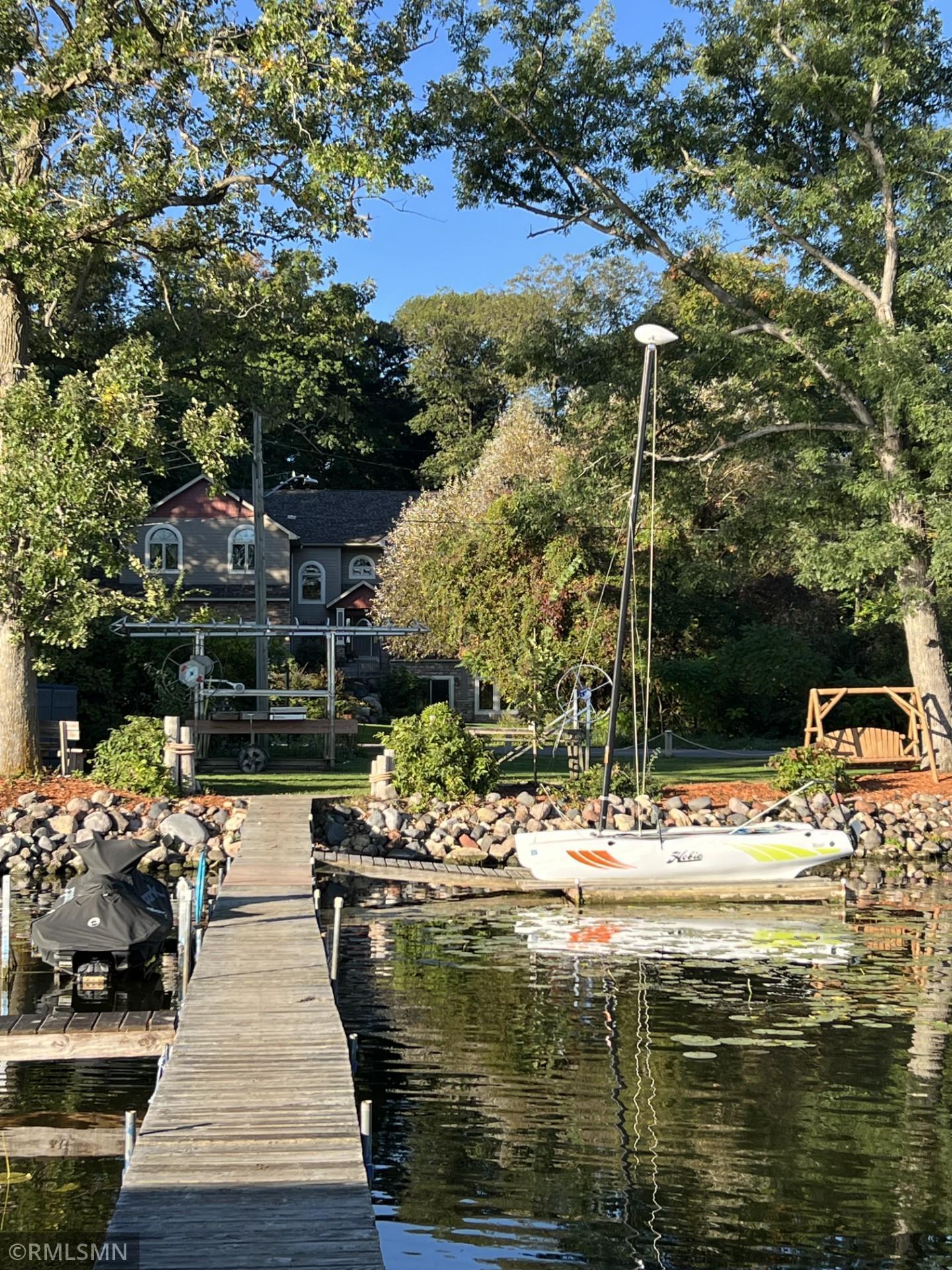 Waterfront home behind mature trees, with a long dock and sailboat on the water.