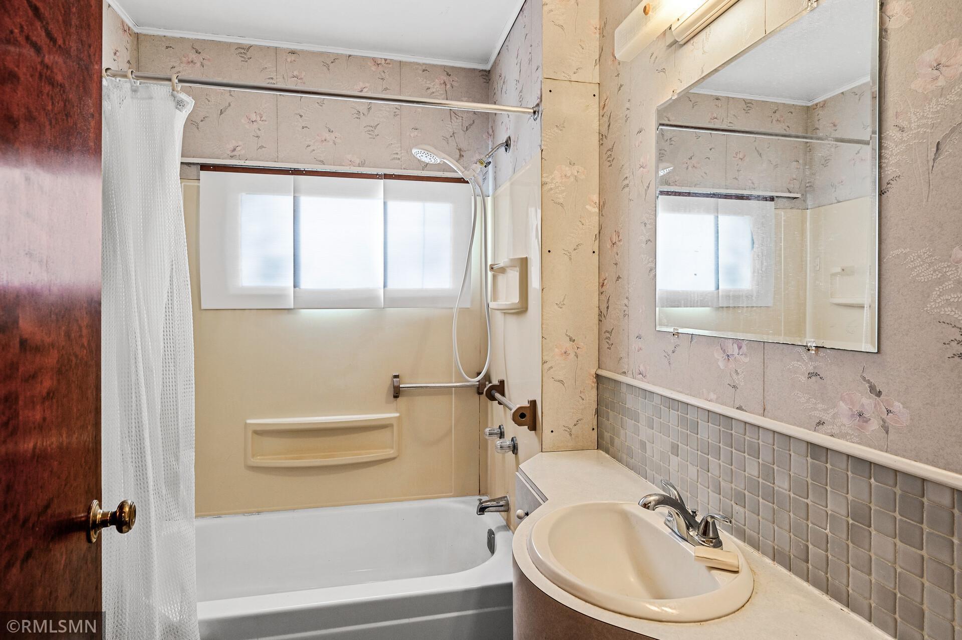 Bathroom with a bathtub-shower, floral wallpaper, tiled wainscoting, and a cream vanity sink.