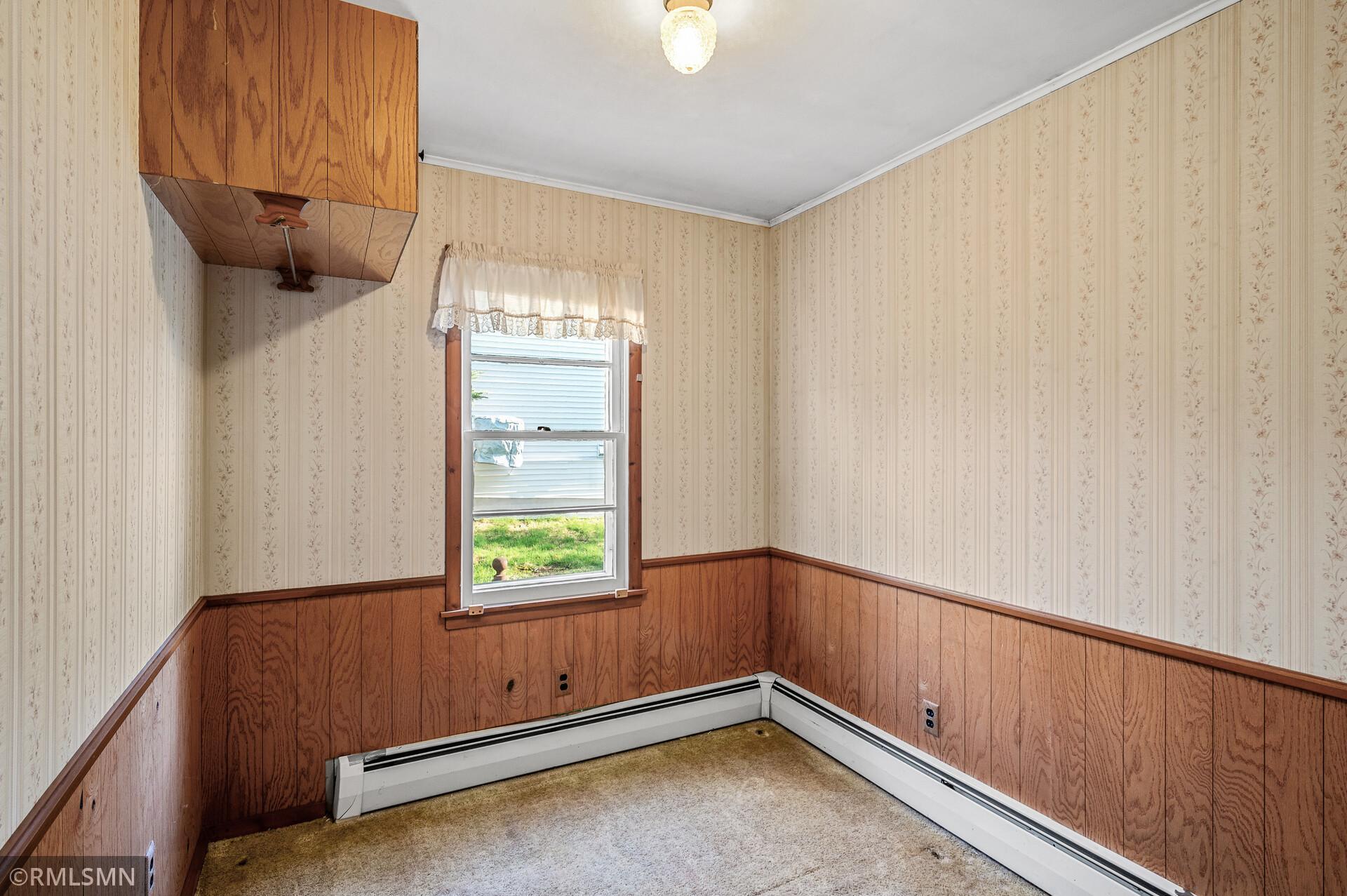 Empty room with floral wallpaper, wood-paneled walls, a lace-curtained window, and baseboard heaters.