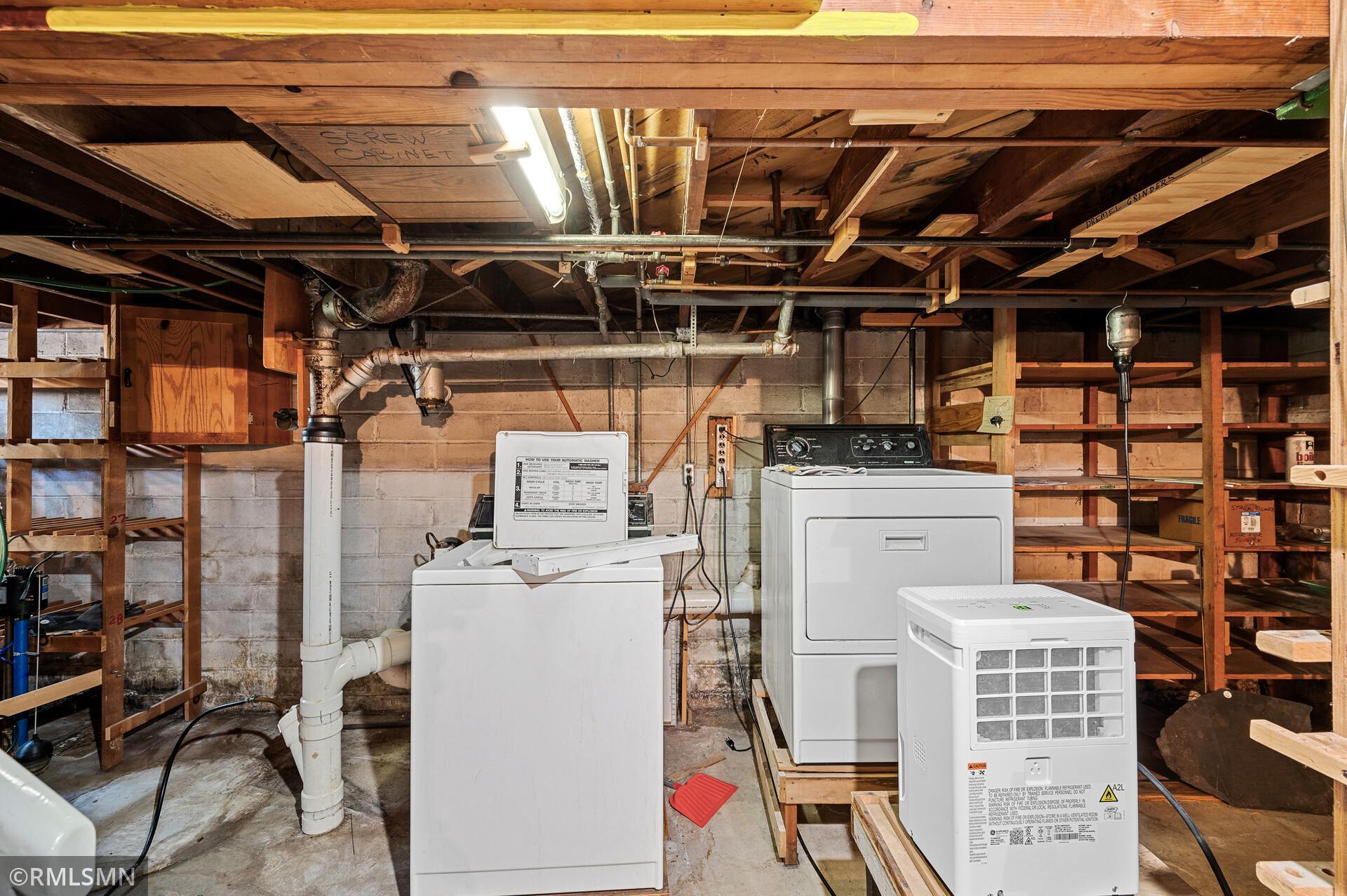 Basement laundry area with washer and dryer beneath exposed joists, surrounded by wooden shelving.