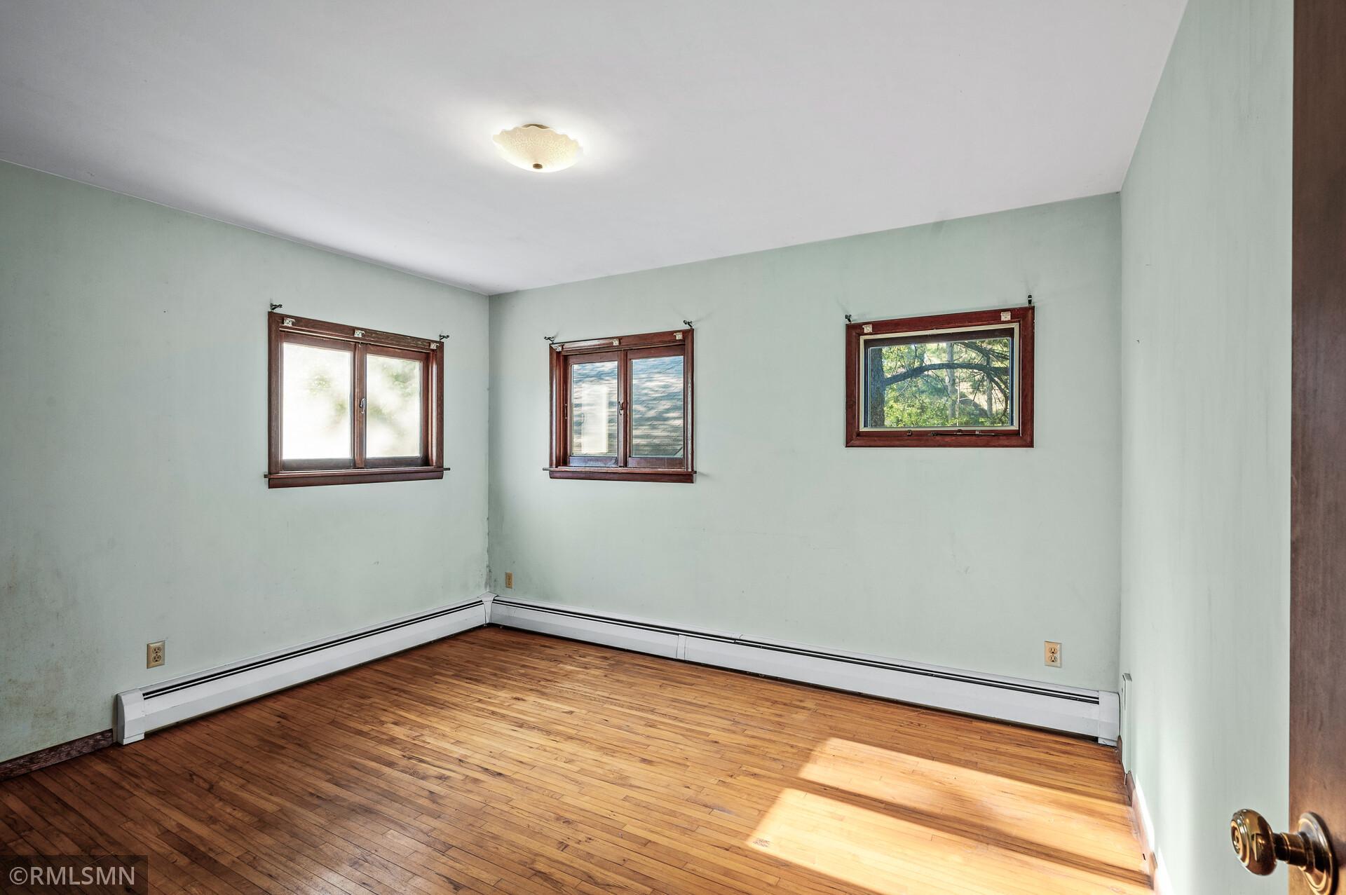 Empty room with hardwood floors, pale green walls, three windows, and baseboard heaters.