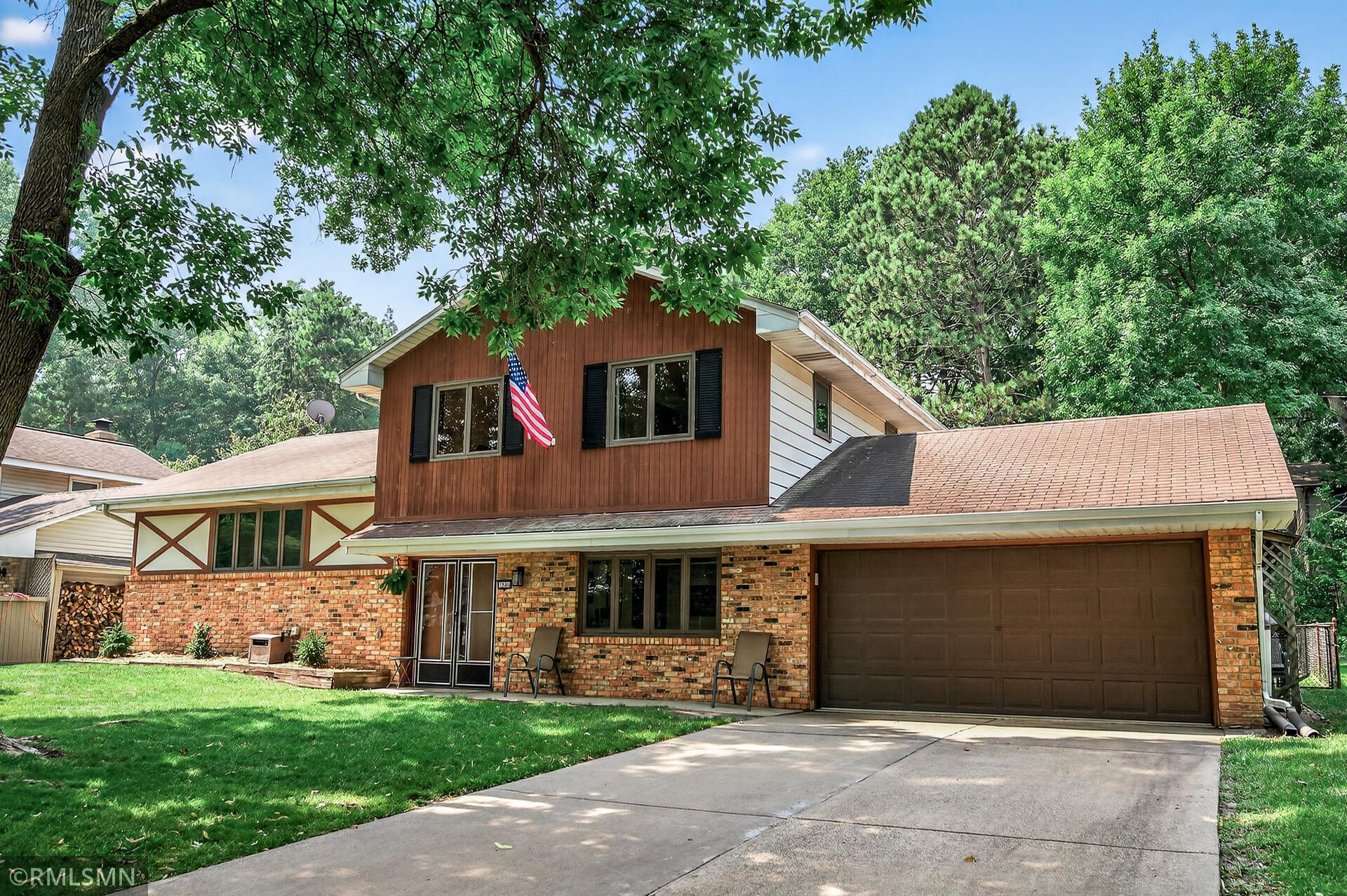 Two-story brick and wood-sided home with an attached garage, American flag, and mature trees.