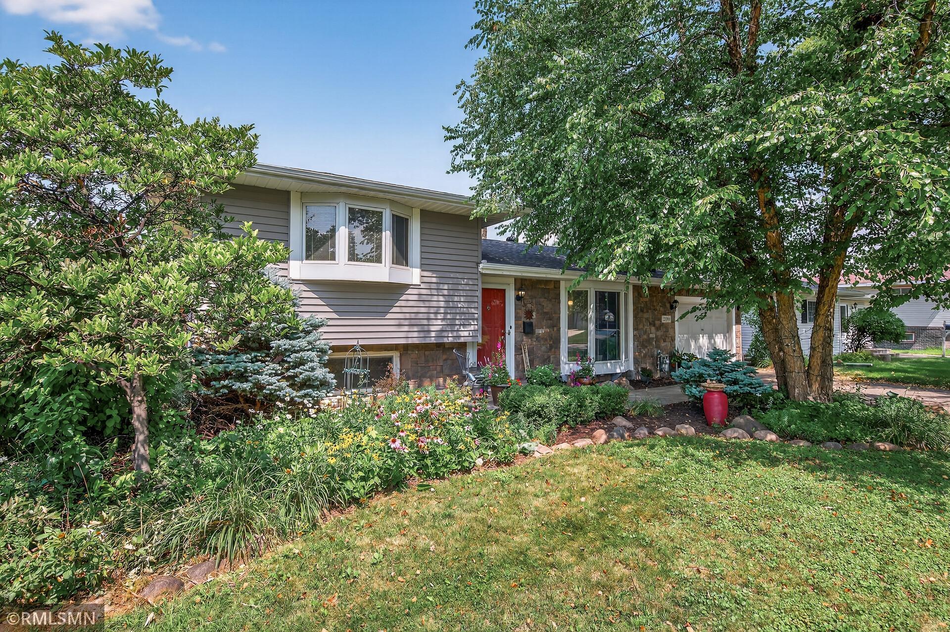Split-level house with a red front door, mature trees, and a colorful flower garden.