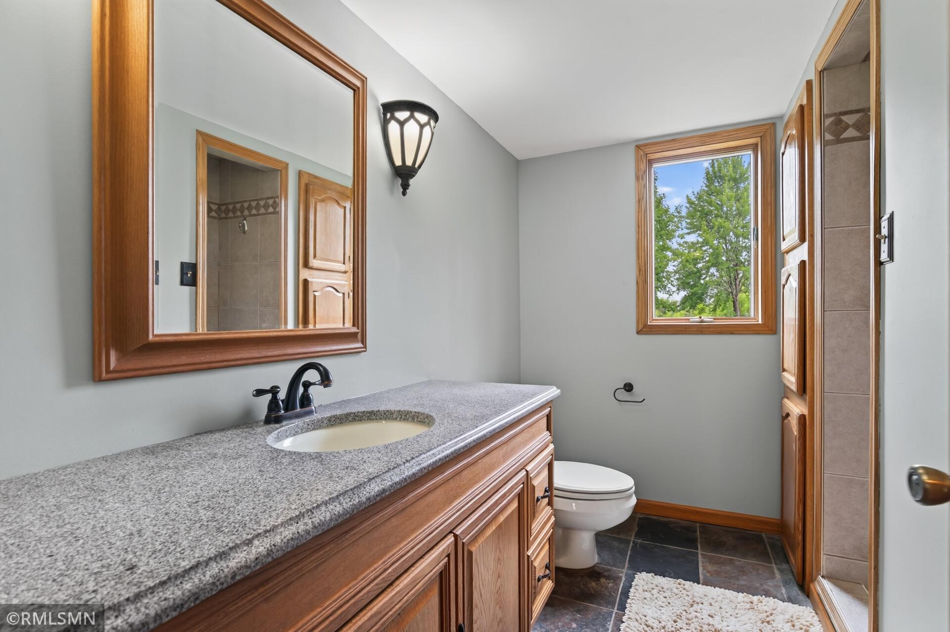 Bathroom with a wood vanity, speckled countertop, large mirror, and window overlooking trees.