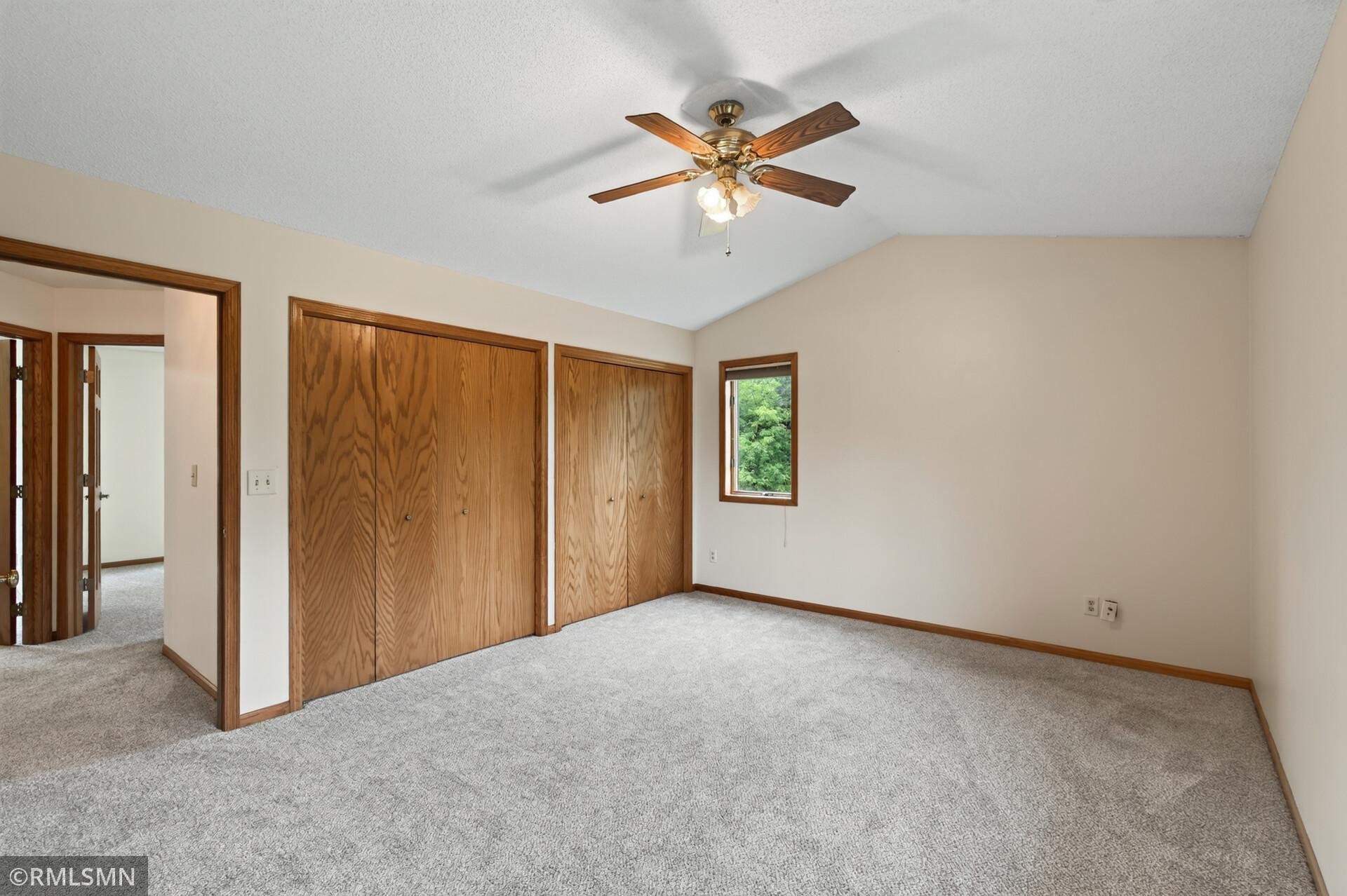 Empty carpeted bedroom with wooden closet doors, a ceiling fan, and a small window.